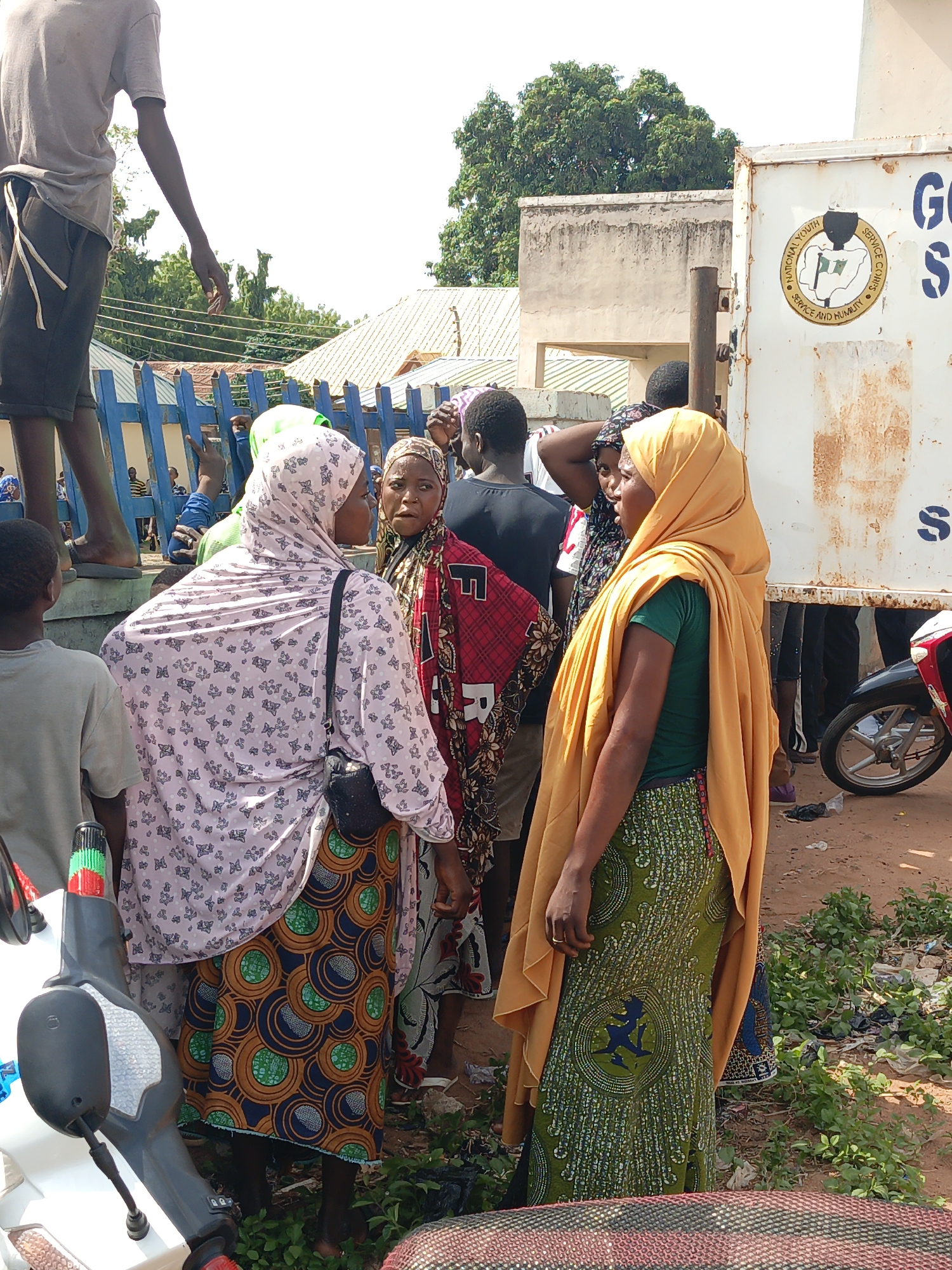The image captures a scene in Lafia, Nigeria, presenting a group of people standing in what appears to be a public area. Three women are prominent in the foreground. One is wearing a pink hijab with a floral pattern and a colorful patterned skirt, another has a red and black patterned wrap, and the third wears a green top, a patterned green skirt, and a yellow head covering. They are interacting with each other. Other individuals are also present, including a boy standing on a blue fence and several people near a white truck with the logo of the National Youth Service Corps. A motorcycle is parked near the women. The ground is a mix of dirt and vegetation. Buildings are visible in the background.