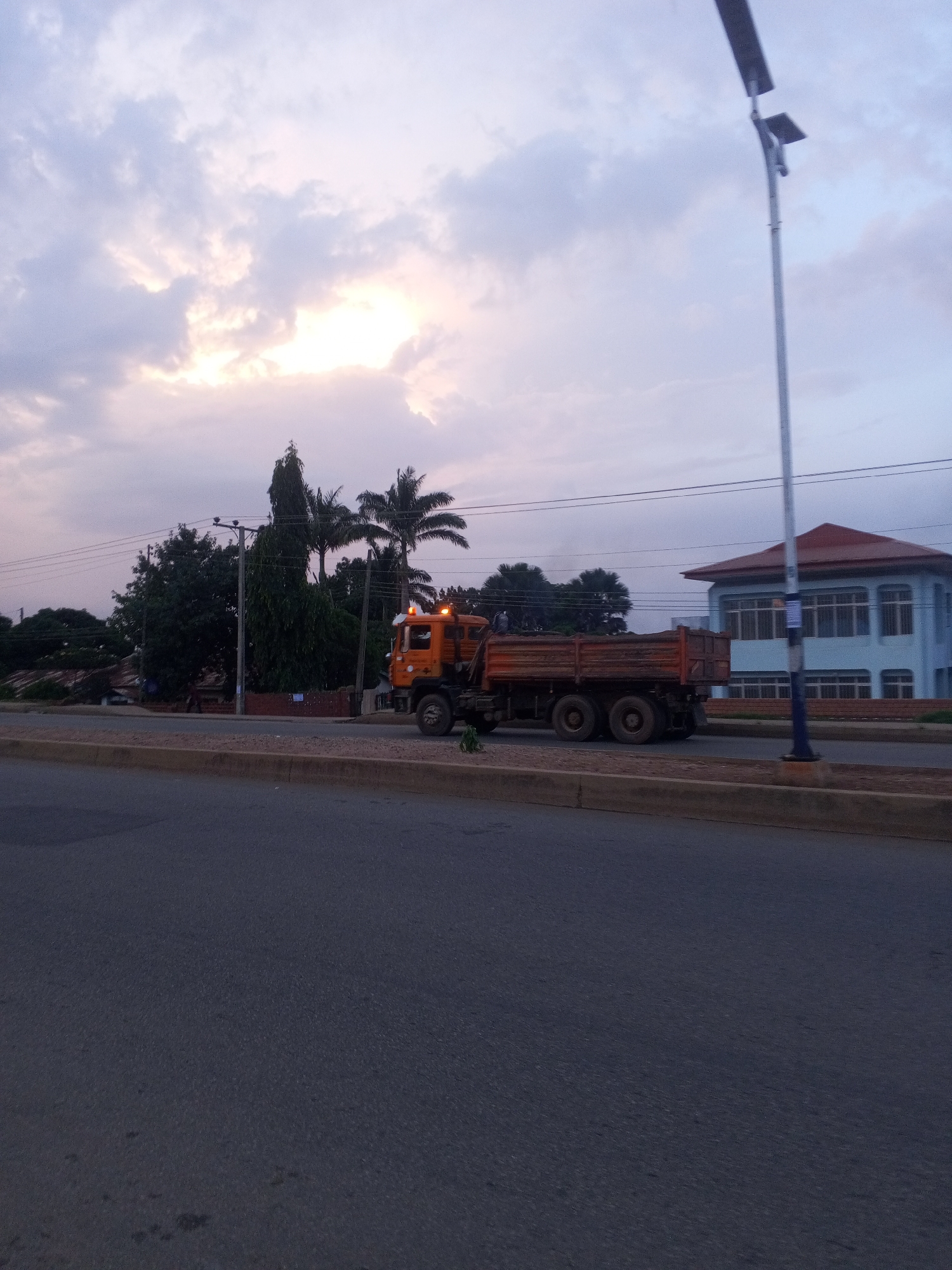 An orange dump truck is parked on the side of a road in Bukuru, Nigeria, with its bed filled with dirt. The sky is overcast with some sunlight peeking through the clouds. In the background, there are palm trees, a blue building with a red roof, and a street light. The road appears to be paved, with a concrete median.