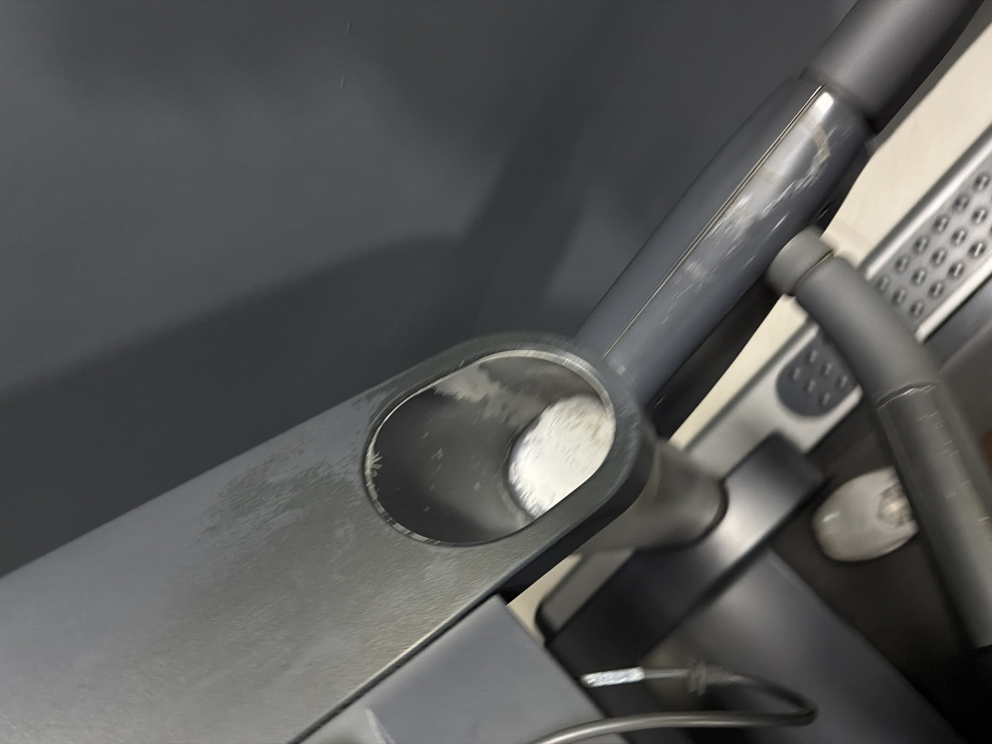 The image shows a close-up, overhead view of a grey treadmill console with a cup holder containing a white powdery substance. The cup holder is circular and appears to be made of hard plastic. The substance inside is fluffy and white, resembling powdered sugar or a similar fine powder. The treadmill console has some scuff marks and a slightly worn appearance. In the background, a dark grey wall is visible, and a portion of a person's shoe and leg is blurred in the lower left of the frame, suggesting the treadmill is in use. The lighting is overhead and slightly diffused. The image does not provide any information or visual cues that would place it in Cancun, Mexico.