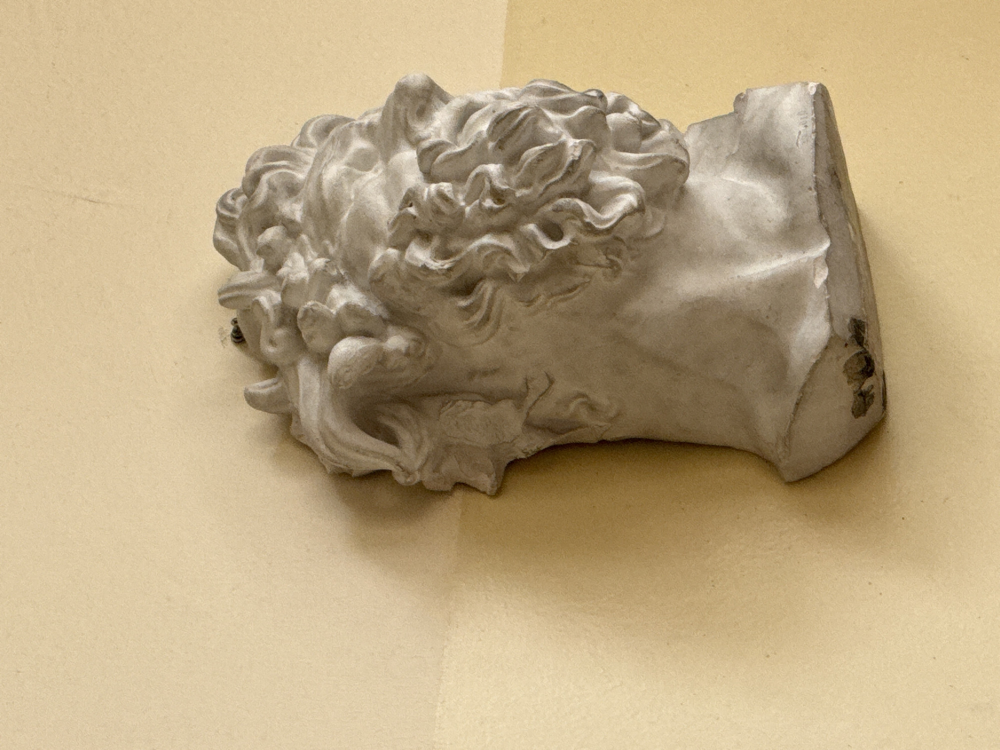 A plaster bust of a classical figure, possibly a representation of a deity or a mythological character, is mounted on a wall at an angle, within a corner of a building in Yerevan, Armenia. The bust is off-white and appears weathered, with some areas of the back seemingly broken off. The wall behind the bust is a plain, light yellow or beige color. The bust is the main subject of the image, taking up a significant portion of the frame. The surrounding wall provides context.