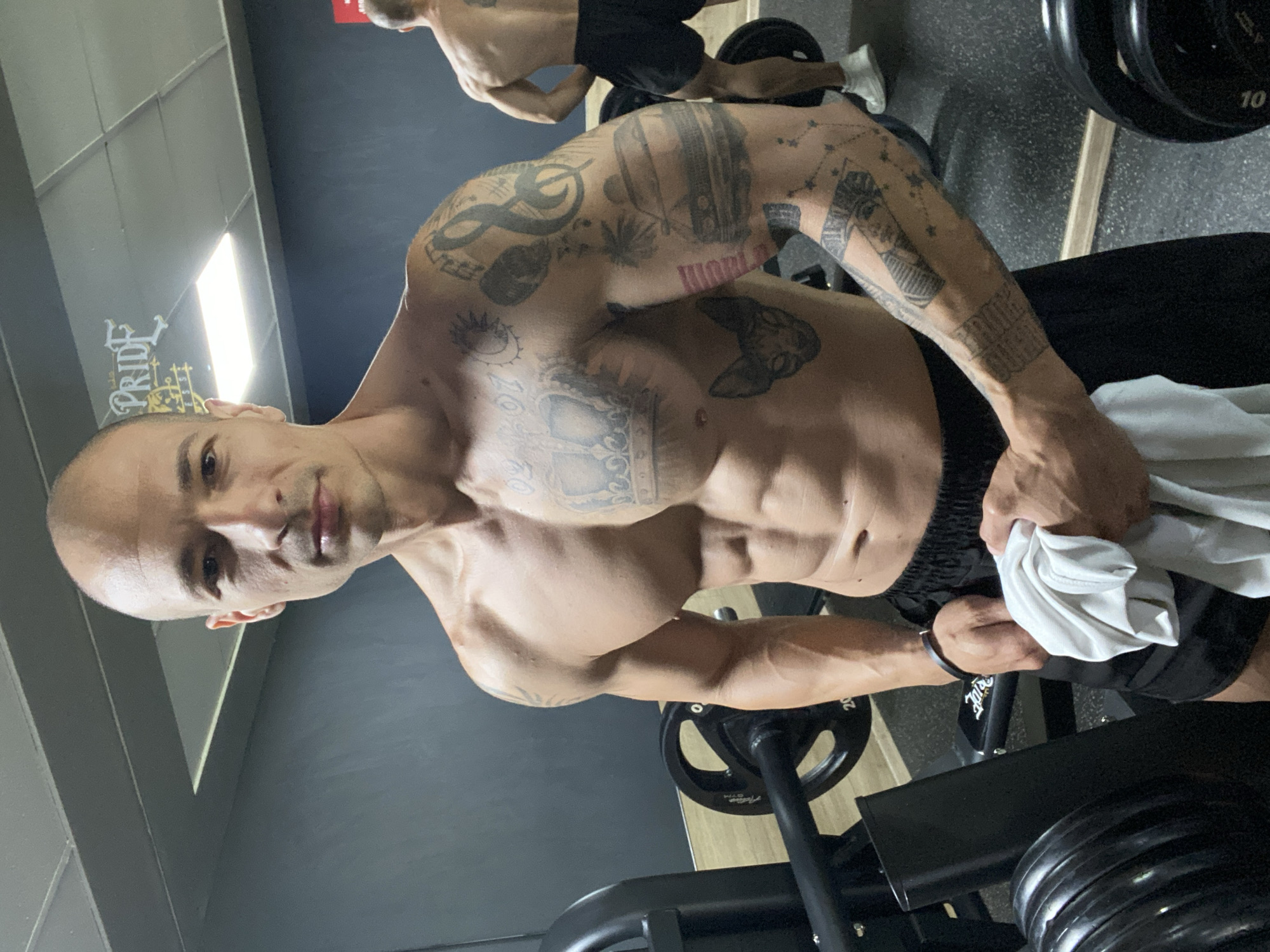 A man with a shaved head, numerous tattoos on his upper body, and toned abdominal muscles is standing in what appears to be a gym, possibly in Pleven, Bulgaria, based on clues in the environment. He's holding a white towel and seems to be wiping sweat. The background features gym equipment, including weight plates and a leg press machine. There's another partially visible person in the background, also shirtless. The gym has grey walls, a bright light fixture, and possible signage.
