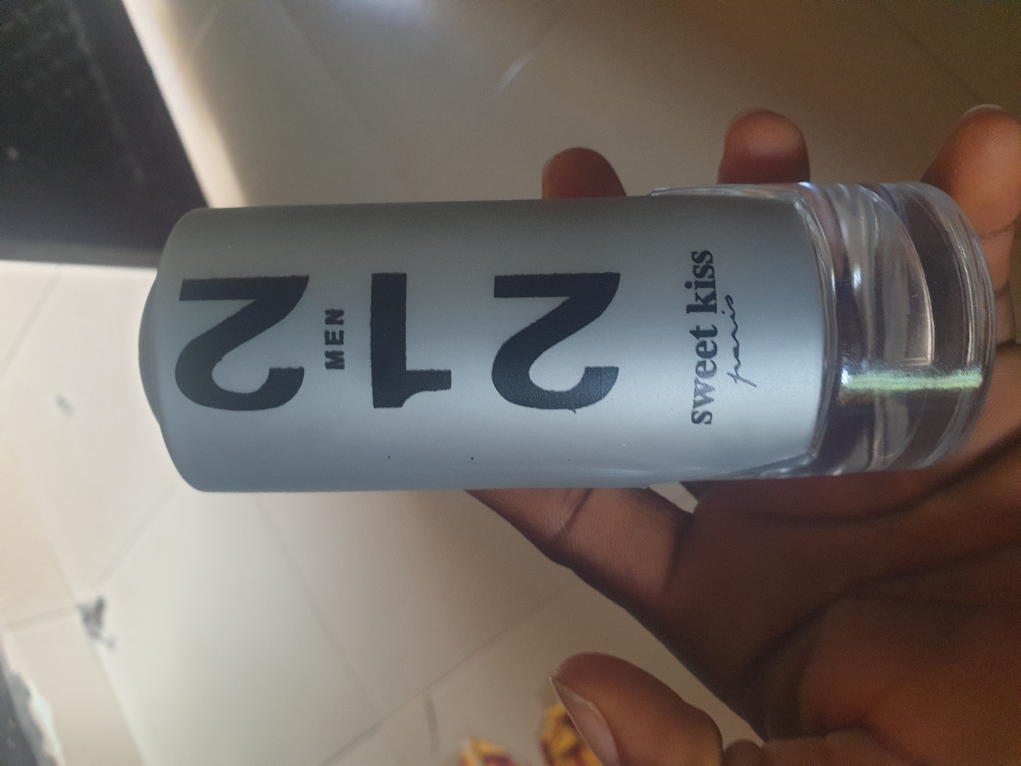 A person's hand is holding a silver bottle of perfume. The bottle has black text on it that reads "212 MEN" and "sweet kiss paris". The background is a light-colored tiled floor.