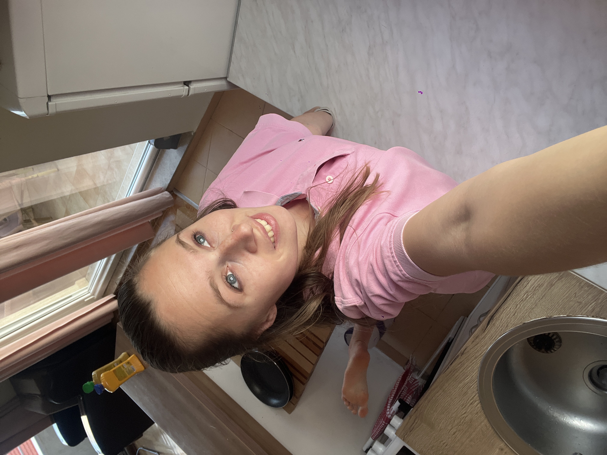 The image shows a woman in a pink polo shirt standing in a kitchen. She is smiling and looking at the camera. The kitchen has light-colored cabinets, a stainless steel sink, and a tile floor. A frying pan rests on a wooden cutting board next to the sink. The window in the background gives the impression that this is likely Boreti, Montenegro.
