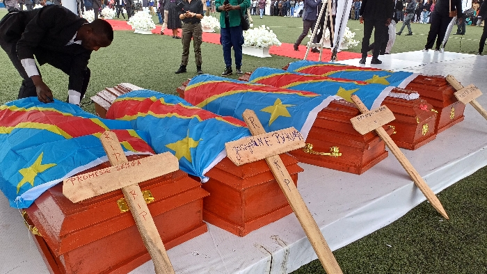 A public memorial service is depicted outdoors on the artificial turf of the Stade de l'Unité in Goma, Democratic Republic of the Congo. In the mid-ground, six small, reddish-brown wooden coffins are arranged in a linear row on a white-draped platform. Each coffin is covered with the flag of the Democratic Republic of the Congo. Directly in front of each coffin, a simple, light-colored wooden cross is staked into the ground, bearing handwritten names such as "PROMESE SENGA" and "GREGANE TUYIVA," along with "R.I.P." written in red marker. In the foreground, a man in a dark suit with a white shirt is bent over the leftmost coffin, appearing to adjust the flag or an item on the coffin. In the background, numerous individuals, some in formal attire, are visible standing and walking on the green artificial turf and a red carpet. White floral arrangements on white pedestals are positioned along the red carpet, indicating a ceremonial setup. The lighting is natural and diffused.