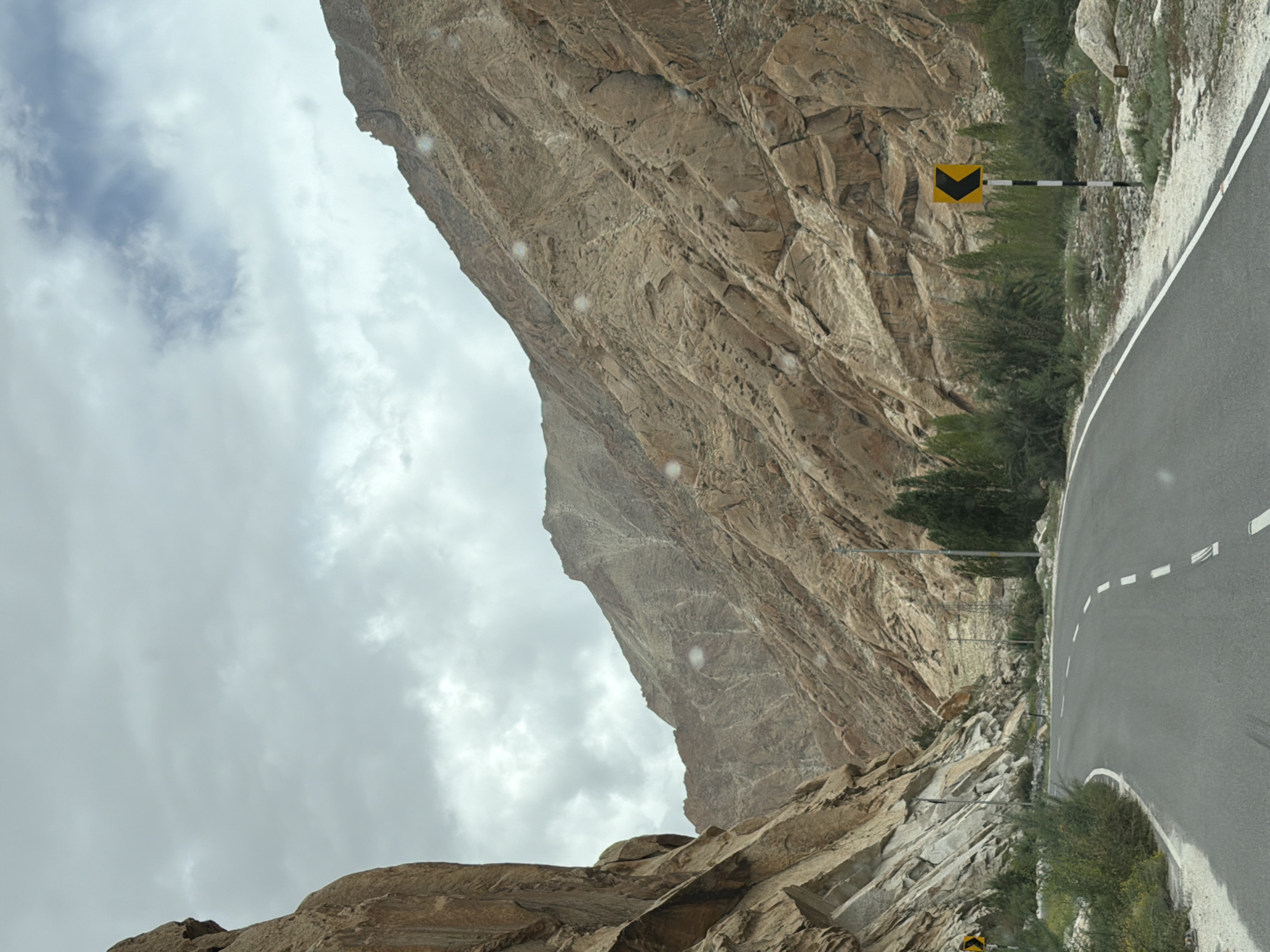 This is a photograph taken from a vehicle traveling on a road through a mountainous region. The road curves to the left and is paved, with white dashed lines marking the lanes. On the left side of the road, a steep, rocky mountain face dominates the scene, composed of beige and gray rock formations. On the right side of the road, another, less steep mountain slope rises, also covered in rugged, textured rock. The upper portions of the mountains are partially obscured by a cloudy sky. Vegetation, including green bushes and trees, grows along the sides of the road and at the base of the mountains. A yellow traffic sign with a black arrow pointing left is visible on the right side of the road, mounted on a striped pole. Power lines are also visible running along the mountainside. The image appears to have been taken during daylight. The location is the city of Likche, India.