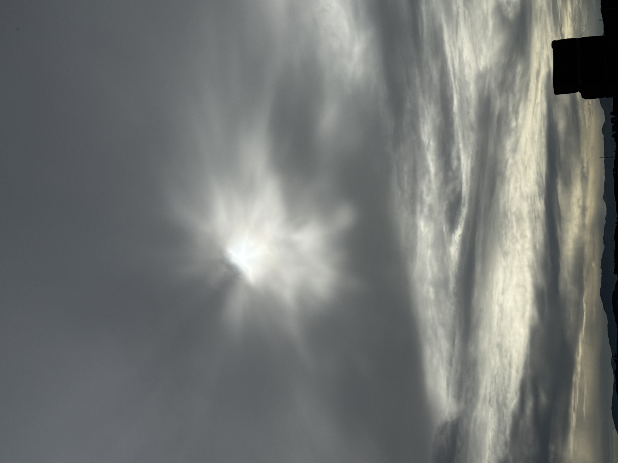 A vertical, medium-shot photograph captures a dramatic sky filled with dark, ominous clouds. The sun, partially obscured by the clouds, shines through with brilliant rays, creating a halo-like effect in the upper-center of the frame. The clouds are layered, with the lower layers appearing lighter and more textured, hinting at a dramatic weather event. In the bottom right corner, a dark, silhouetted structure, appearing to be two stacked rectangular blocks, stands in stark contrast to the bright sky. The horizon line at the bottom of the image shows a faint silhouette of distant hills or mountains. The overall mood is somber and awe-inspiring, with a focus on the power and beauty of nature. The scene is set in Unknown, Unknown.
