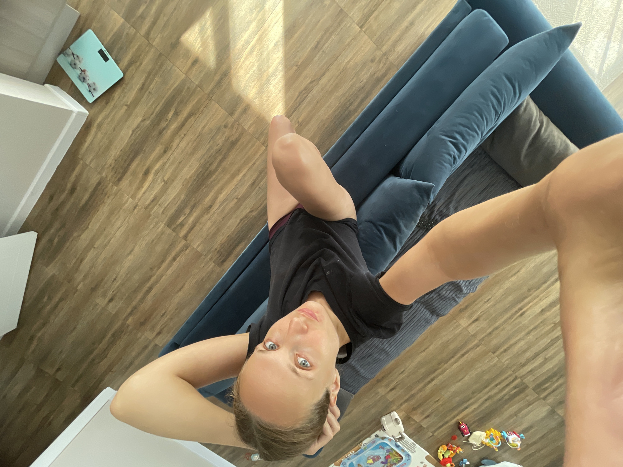 A high-angle, top-down selfie is taken by a woman lying on a long, blue sectional sofa. She has light skin, blue eyes, and light brown hair, which is pulled back. She is wearing a black t-shirt and dark shorts. Her right arm is bent, with her hand resting on her head, and her left arm is also bent, with her hand on her head as well. She is looking directly at the camera with a neutral expression.
The sofa is positioned on a wooden floor with a plank pattern. Sunlight streams in from the right, casting distinct shadows on the floor and the sofa. To the left of the sofa, there's a white piece of furniture. In the foreground, on the floor, there is a children's water play mat with various colorful toys scattered around it, including what appears to be a small toy car and other figures. Further away, near the wall, a light blue digital scale with an image of cotton branches is visible.
The overall scene suggests a relaxed indoor setting, likely a living room, possibly during daylight hours due to the strong sunlight. The presence of children's toys hints at a family environment. The location context provided is Podshivalovo, Russia. There is no visible text in the image, other than the digits on the digital scale which are not legible.