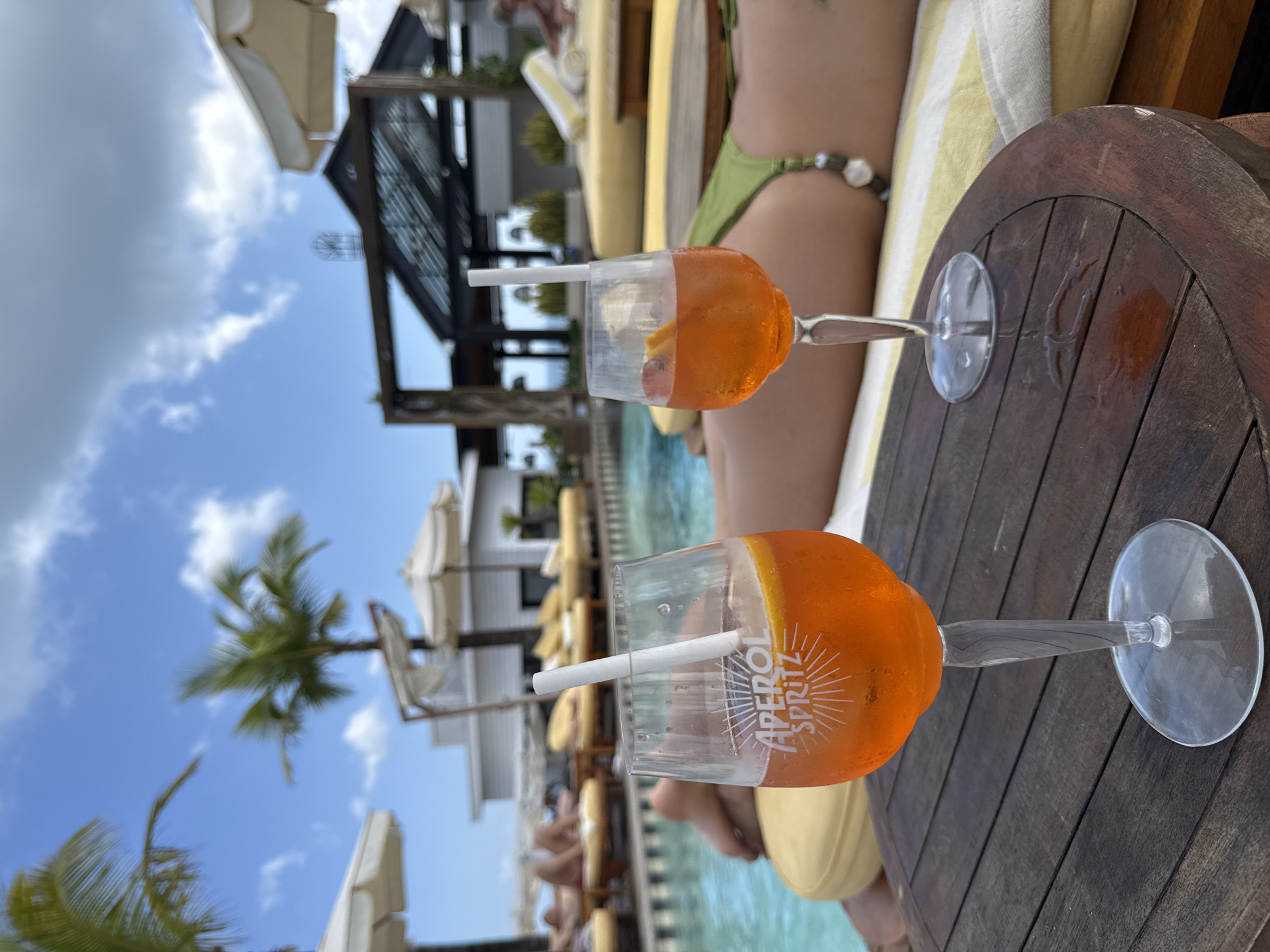 The image shows two Aperol spritz cocktails on a wooden table, likely in an outdoor setting. In the background is a swimming pool, lounge chairs with yellow cushions, and beach umbrellas, indicating a resort or upscale beach club. People are lounging by the pool, suggesting a relaxed atmosphere. There is a structure with a black pergola or trellis, possibly a bar or restaurant area. In the background are tropical trees, including palm trees, and a blue sky with clouds.
Based on the available information, and the presence of Aperol Spritz drinks, beach loungers and umbrellas, and tropical landscape, a possible location in North Kuta, Indonesia could be a luxury beach club or resort.