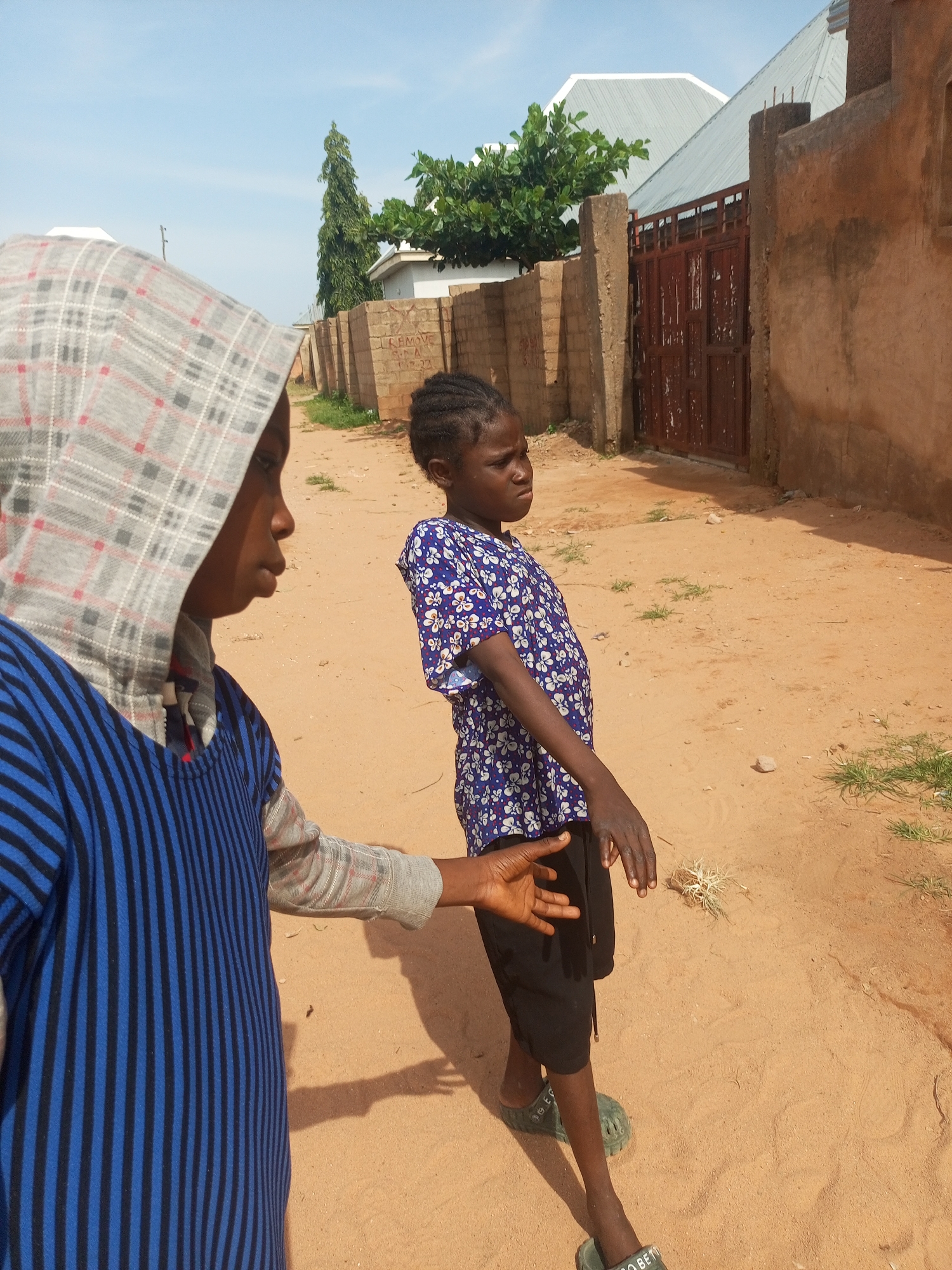 Two young children are standing on a sandy dirt road in Tempure, Nigeria. The child on the left, facing the right, is wearing a grey hooded sweatshirt with a checkered pattern and a blue and black striped shirt underneath. The child on the right, facing left, is wearing a blue short-sleeved shirt with a white floral pattern and black shorts. The child on the right has their right arm extended forward, with their hand open. The environment includes a brown plastered wall on the right with a red metal gate. Further in the background, there are brick walls, trees, and buildings with corrugated metal roofs. The ground is covered in light brown sand with sparse patches of dry grass. The sky is clear and blue with a few wispy clouds.