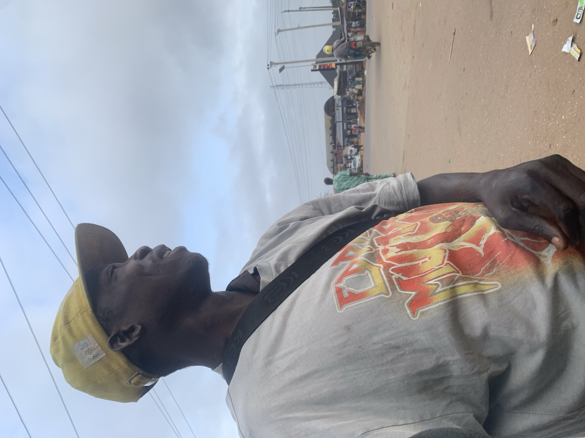The image shows a medium shot of a dark-skinned man wearing a yellow baseball cap and a light gray t-shirt with a graphic design. He is positioned in the foreground, looking to his right. The t-shirt has a large, colorful design on the chest that appears to be an image with flames. A dark strap, possibly from a bag, is visible across his chest.
The background depicts a street scene in Shagamu, Nigeria, under an overcast sky. Several power lines stretch across the upper portion of the frame. In the middle ground, there are buildings that appear to be shops or market stalls. Several people are visible, some walking and others possibly interacting in front of the establishments. A person on a motorcycle, wearing a yellow helmet and a gray jacket, is moving away from the camera on the right side of the street. Cars and other motorcycles are also present on the street, suggesting a moderately busy urban environment. The road surface is paved and appears to be a light brown color, with some scattered debris.