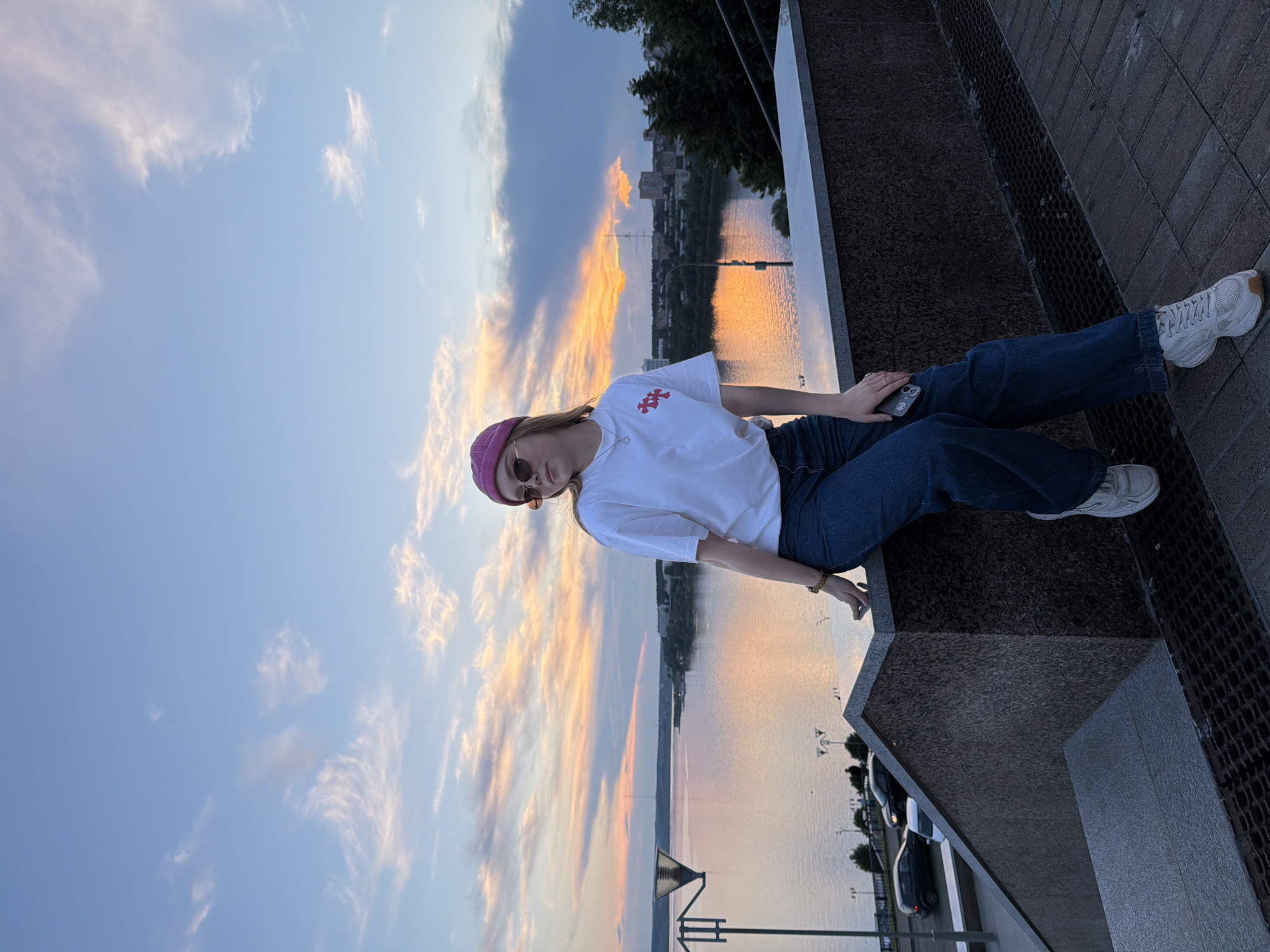A young woman wearing a pink beanie, sunglasses, a white t-shirt with a red emblem, and dark blue jeans is sitting on a granite ledge overlooking a wide river at sunset. The sky is filled with streaks of orange, pink, and blue clouds. In the background, a city skyline with buildings and trees is visible across the river. Cars are parked on a street below the ledge. The woman is holding a smartphone in her right hand. The scene appears to be the embankment of the Izh River in Izhevsk, Russia, likely near the Friendship of Peoples monument area.