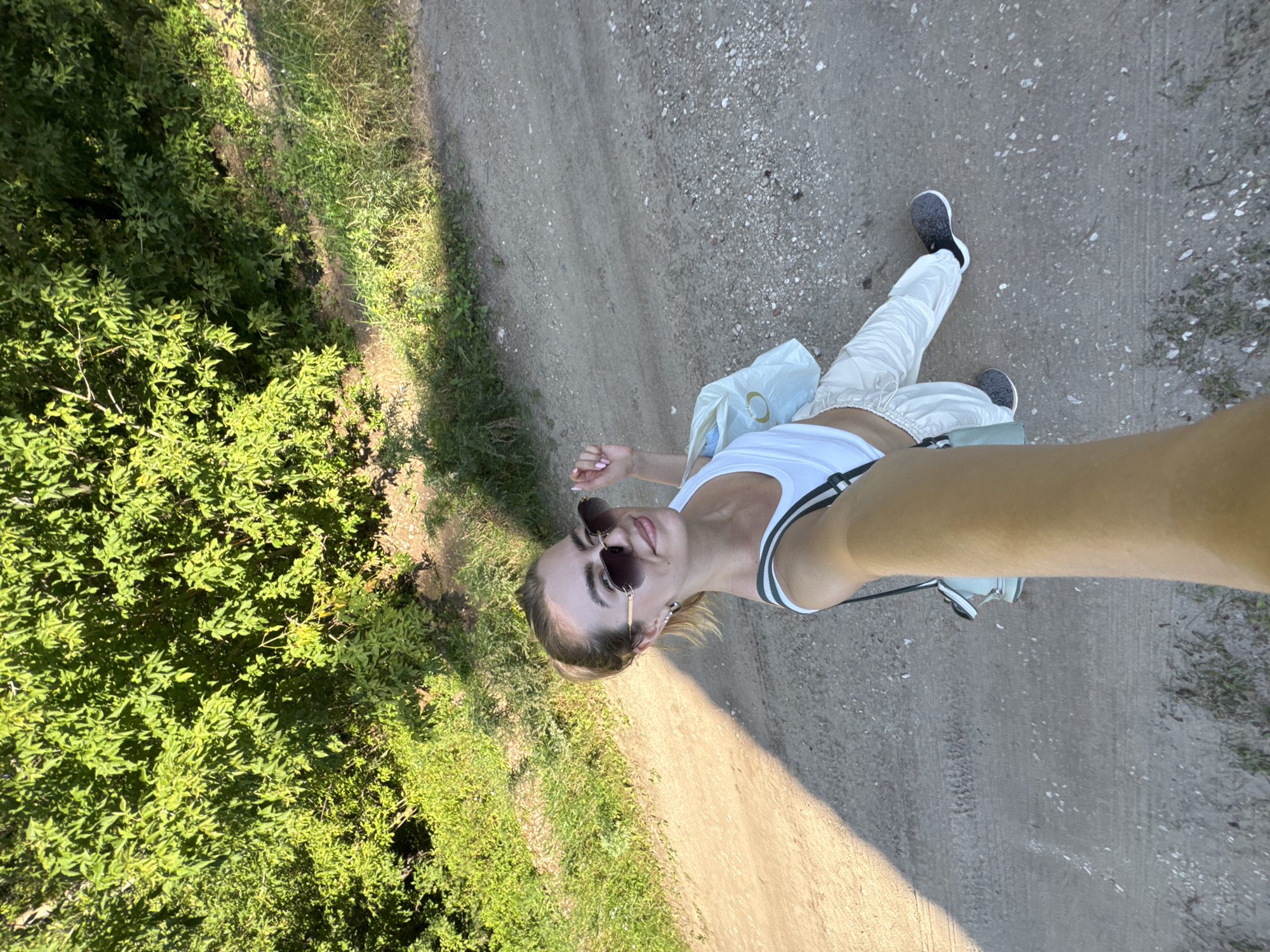 A young Caucasian woman is taking a selfie outdoors on a dirt path. She is wearing a white tank top, light-colored pants, and sunglasses. A light blue tote bag is slung over her shoulder. She has her hair pulled back in a ponytail. The background consists of green foliage and a dirt path, with dappled sunlight casting shadows. The location is not specified beyond "Unknown, Unknown."