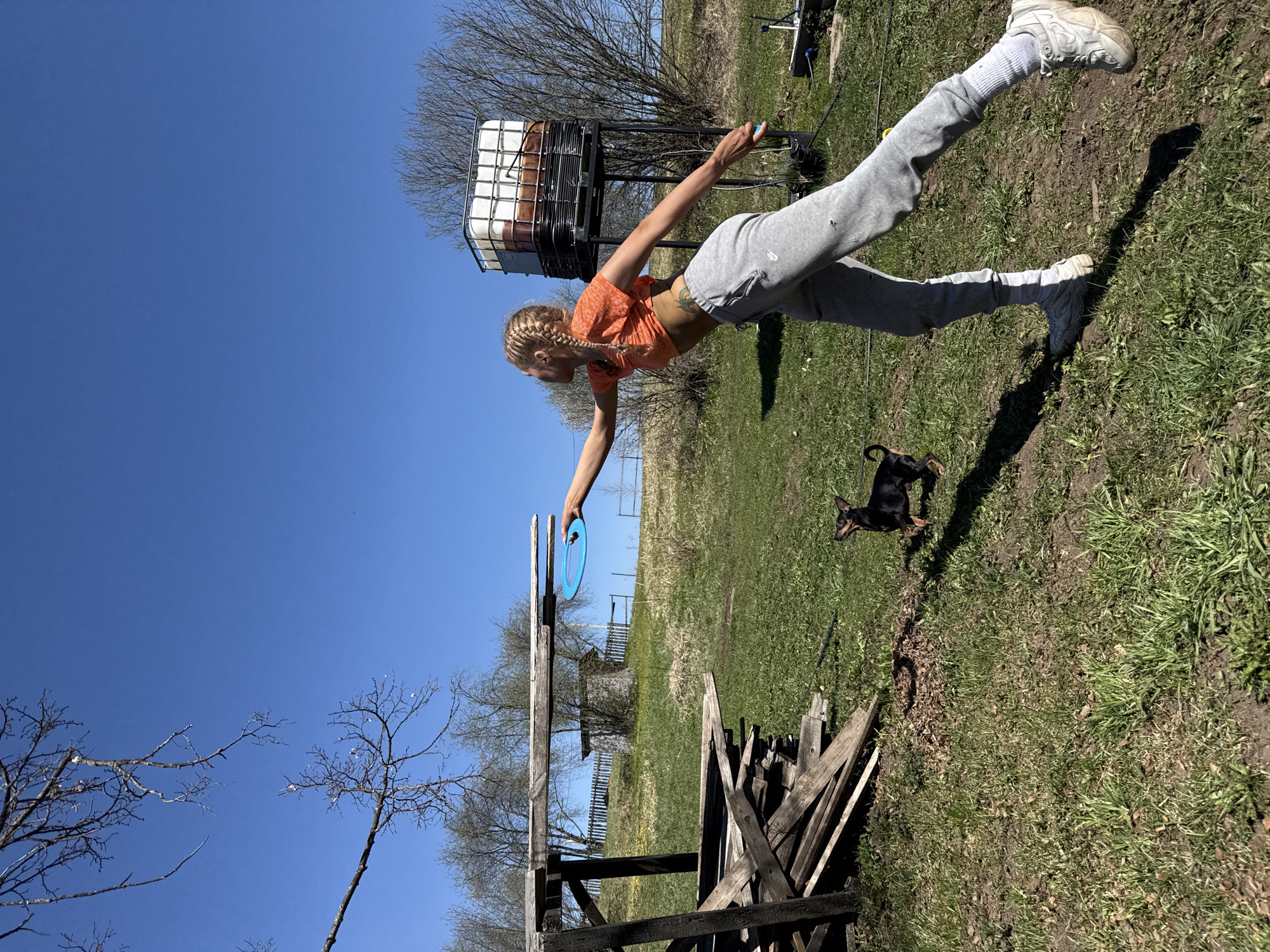 A young woman with blonde braids is standing outdoors on a grassy field. She is wearing an orange crop top, grey sweatpants, white socks, and white sneakers. Her arms are outstretched, holding a blue ring. To her right, a small black and tan dachshund stands on the grass. In the background, there is a water tank on a metal frame, a wooden fence, and a pile of lumber. The sky is clear and blue with a few bare trees visible. The location is likely rural, given the field and lack of urban structures. The city is not visible, and the location cannot be confirmed.