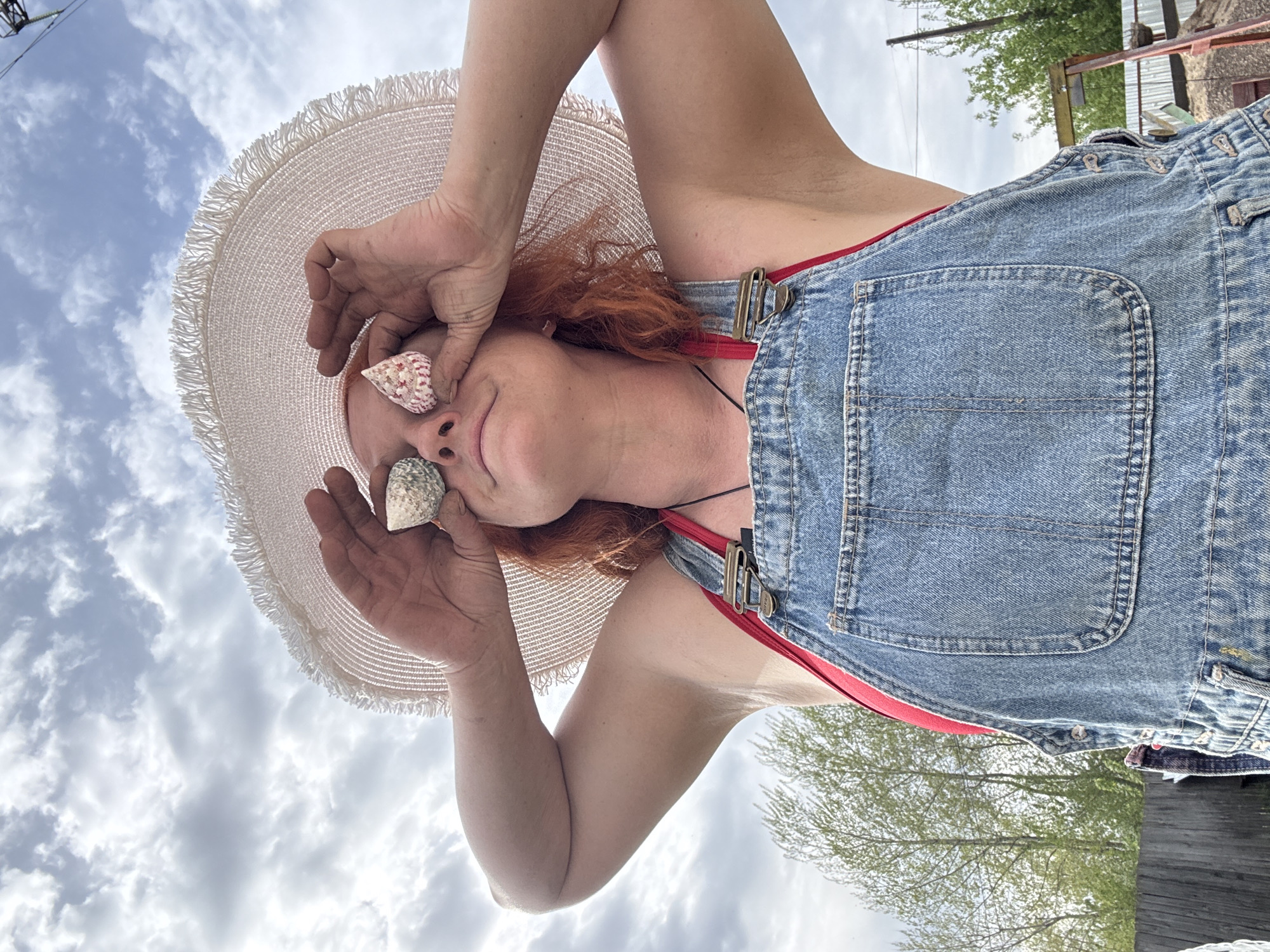 A woman with red hair is lying down, wearing a straw hat, a red top, and blue denim overalls. She is holding seashells to her eyes and nose. The background shows a cloudy sky. The location is Chaikovsky, Russia.