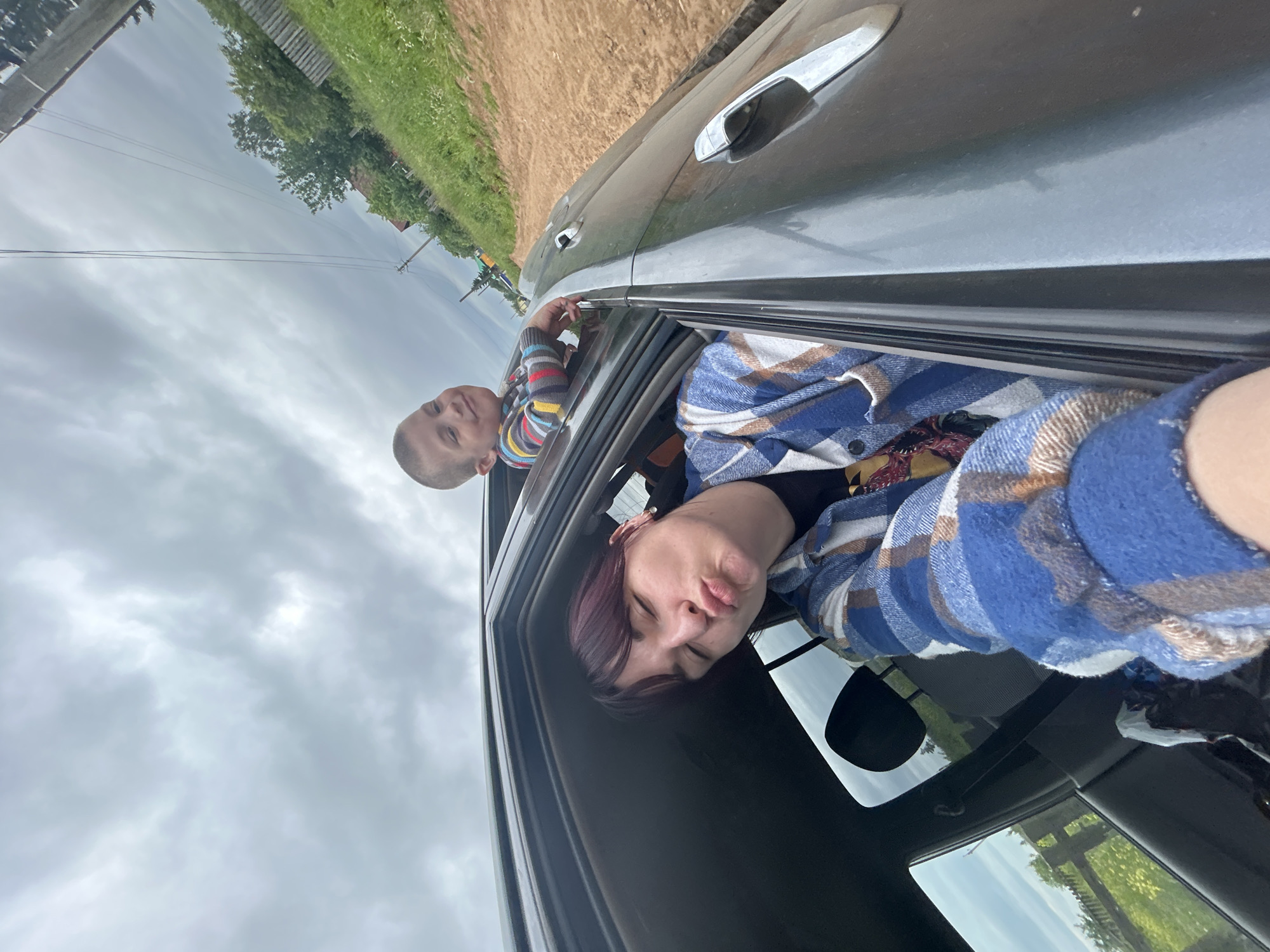 In the city of Foky, Russia, on a cloudy day, a woman with dark hair and a blue and brown plaid shirt is taking a selfie from inside a car, puckering her lips. Next to her, a young boy in a striped shirt leans out of the car window, smiling. The car is on a dirt road with green grass and trees on either side. A wooden fence is visible in the background.