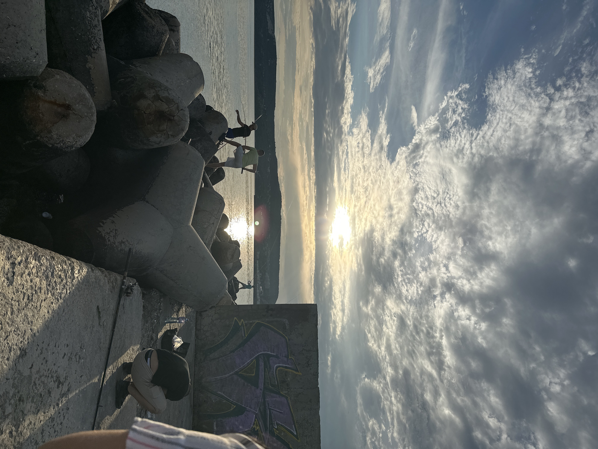 The image captures a coastal scene in Balchik, Bulgaria, during either sunrise or sunset, indicated by the low sun angle and long shadows. The foreground and mid-ground are dominated by a substantial breakwater composed of large, weathered grey concrete tetrapods. Three males are positioned on these tetrapods, actively engaged in fishing in the calm waters of the Black Sea; two are in the mid-ground, one wearing a dark shirt and the other a light green shirt, both holding fishing rods. A third, silhouetted male is visible on a more distant tetrapod, also fishing.
On the right side of the image, a large, vertical concrete wall features prominent purple and yellow graffiti, appearing as a stylized 'V' or similar tag. Adjacent to this wall, on a flat concrete pier, a fourth male, dressed in a black shirt and light shorts, is observed in a deep squat, attending to fishing gear that includes a black tackle box, a plastic water bottle, and plastic bags. A fishing rod rests horizontally on the concrete surface near him. A partial view of a fifth individual, showing a striped shirt, is present in the extreme foreground right, suggesting the perspective of the photographer or a nearby person. The horizon across the water reveals dark, undulating hills or mountains, with subtle indications of a distant settlement. The sky is partially cloudy, displaying various cumulus and stratocumulus formations, and the low sun casts a bright reflection on the water, producing lens flare.
