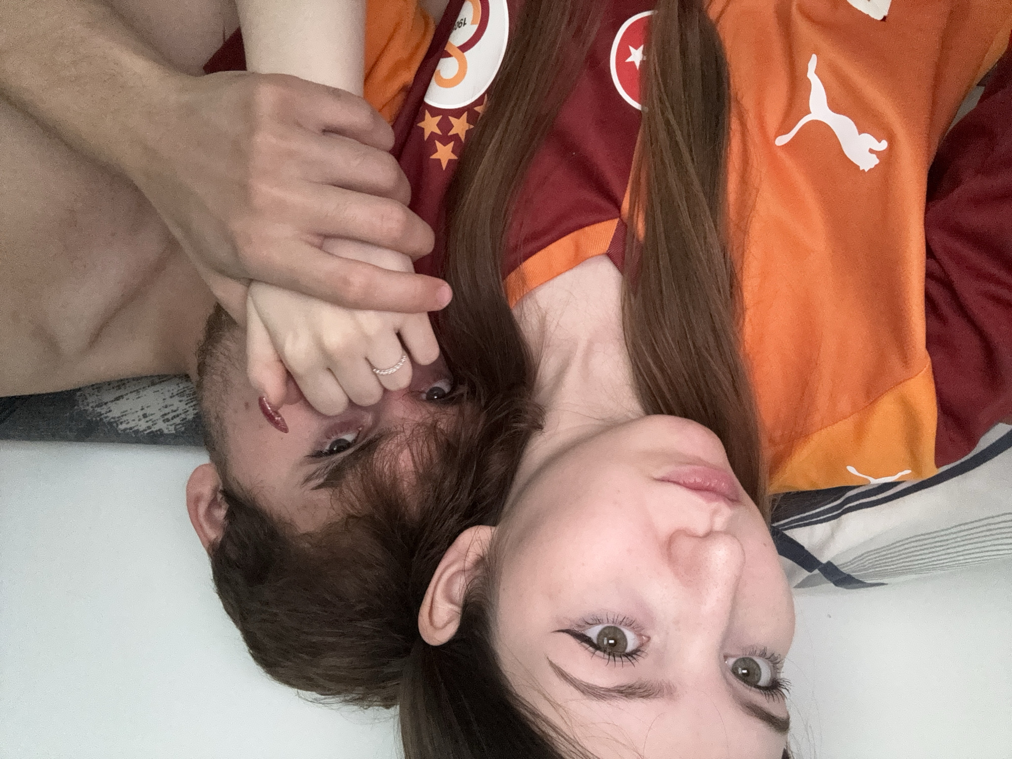 The image shows a close-up, selfie-style shot of a young couple, a male and a female, embracing. The female, on the left, has long brown hair, light skin, and green eyes. She is wearing a maroon and orange Galatasaray soccer jersey with a Puma logo and stars. Her left hand is resting on the male's hand, and she is wearing a ring on her left ring finger. The male, on the right, has brown hair and appears to have a scrape or mark on his cheek. He is holding the female's hand. The background is a plain white wall and a striped pillow. The image does not contain any identifiable landmarks or features that would allow for its location to be determined to a specific city like Ufa, Russia.