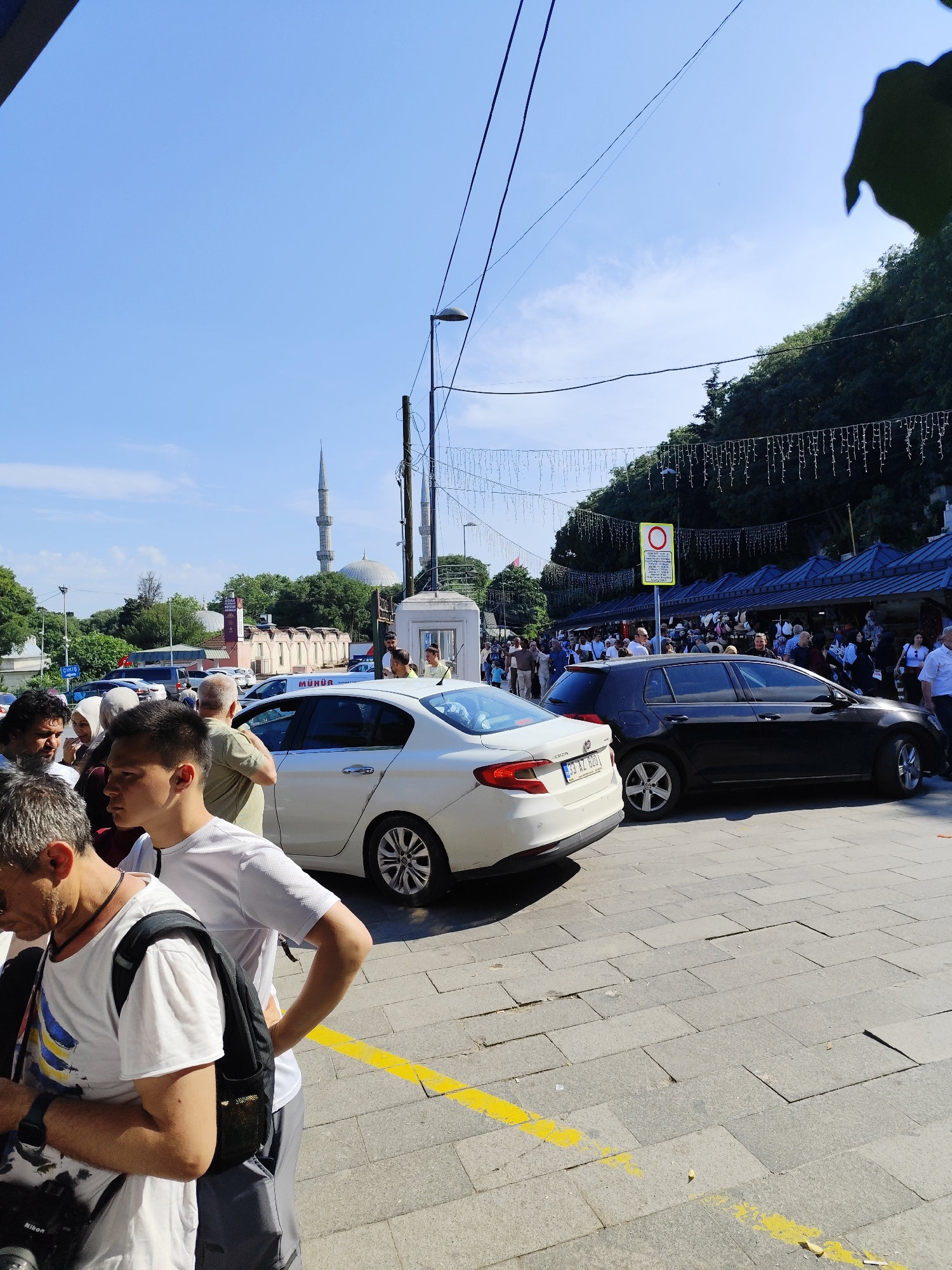 The image depicts a crowded street scene in Eyüpsultan, Turkey, on a sunny day. A white Honda sedan with license plate "33 AZ 826" is parked alongside a black Volkswagen hatchback. Several people are walking or standing near the vehicles, including a man with a backpack in the foreground and a crowd by covered vendor stalls. A mosque with a tall minaret is visible in the background, indicating the location is likely near a religious site. Overhead streetlights and decorative lights extend above the scene. The road is paved with gray rectangular stones and marked with a faded yellow line.