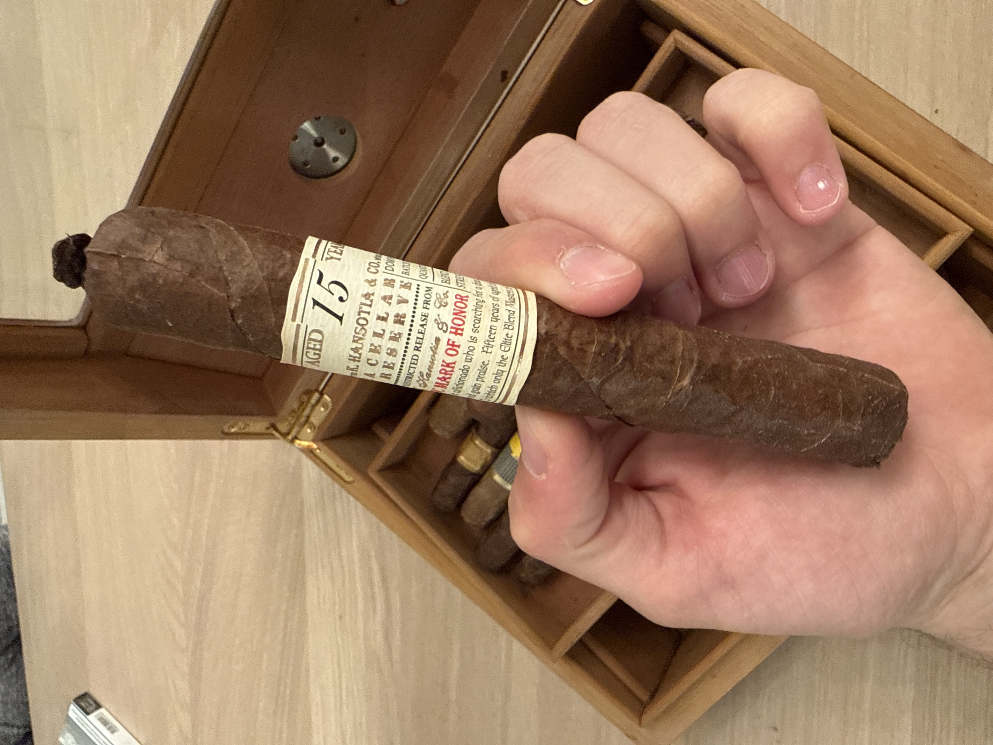 A hand holds a cigar with a label reading "AGED 15 YEARS", "K. HANSOTIA & CO.", "ACELLARB DOM", "RESERVE BATE", "RESTRICTED RELEASE FROM", "Hansotia & BUT", and "MARK OF HONOR STRES". The cigar is brown and slightly wrinkled. The hand is pale with short fingernails. A wooden humidor with a few other cigars is partially visible in the background. The background is a light-colored wooden surface. The location is not specified and cannot be determined from the image.