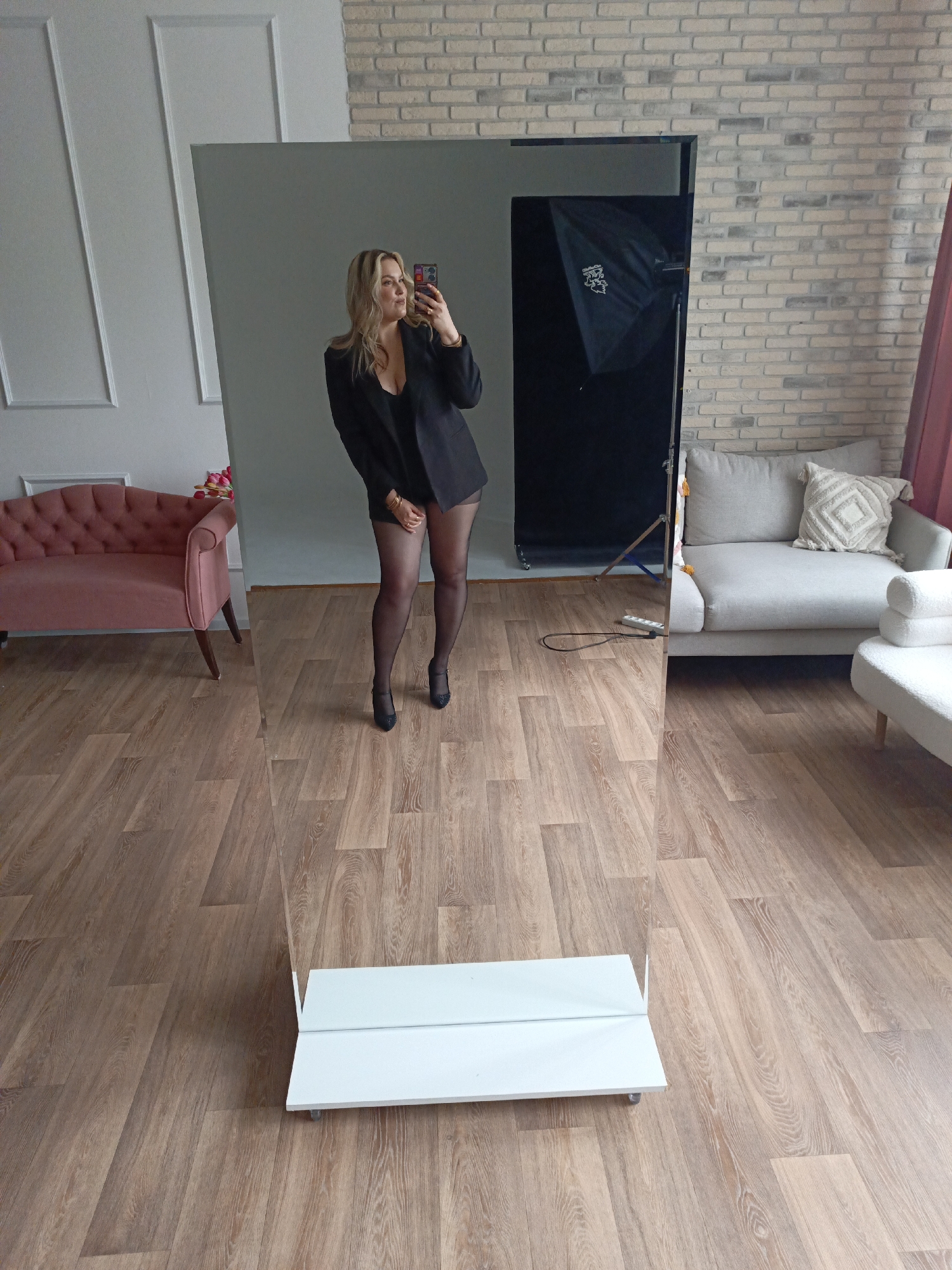 A full-length mirror is positioned on a light brown, wood-patterned floor. Reflected in the mirror is a woman wearing a black blazer, black top, black stockings, and black heels. She is holding a smartphone in her right hand, presumably taking a photo of herself. The background reflected in the mirror includes a light gray wall to the left, a pink tufted couch, and a brick wall to the right with a light gray couch and a lighting umbrella in front of it. The location is Chaikovsky, Russia.