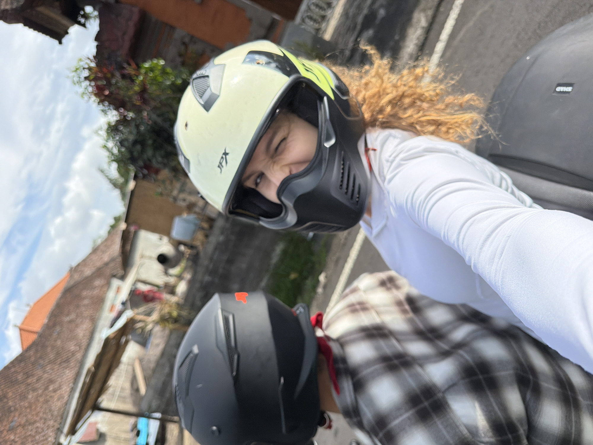 Two individuals, one in front and one in back, are riding a motorcycle on a street in Penebel, Indonesia. The person in the front is wearing a white long-sleeved shirt and a light yellow motorcycle helmet with a black face mask. They are looking at the camera and winking. Their curly, light brown hair is visible. The person in the back is wearing a black helmet and a brown and white plaid shirt with a red bandana tied around their neck. The background shows a residential area with buildings featuring tiled roofs, some with orange tiles. There are trees and vegetation visible, as well as a partially constructed building with cement bags and tools. The sky is partly cloudy.