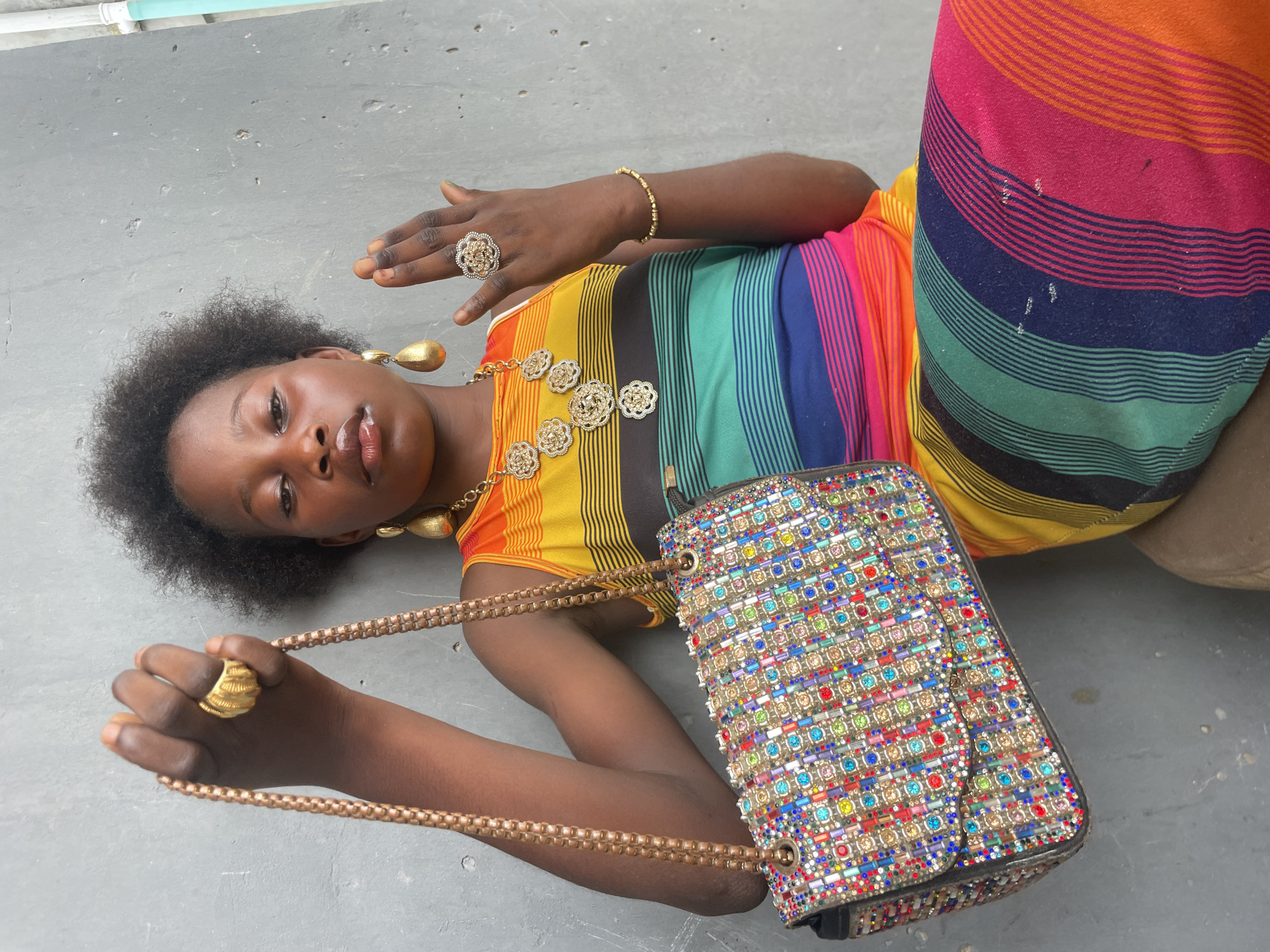A young Black woman is depicted lying on a light gray concrete surface, looking directly at the camera. She has short, natural, curly hair and her lips are slightly parted with a glossy finish. She is wearing a vibrant, multi-colored garment, possibly a dress or top, featuring horizontal stripes in a spectrum of colors including yellow, orange, red, pink, blue, green, and black.
Her attire is accessorized with several pieces of gold-toned jewelry. On her right hand (viewer's left), she wears a large, intricately designed ring with a floral or swirling pattern and a beaded bracelet on her wrist. On her left hand (viewer's right), she wears a large, ribbed gold-toned ring and is holding the chain strap of a handbag. Her neck is adorned with a gold-toned necklace featuring multiple circular medallions, and she wears large, teardrop-shaped gold-toned earrings.
The handbag, positioned beside her, is a prominent feature. It is rectangular and heavily embellished with a mosaic-like pattern of numerous colorful rhinestones or beads in shades of red, blue, green, yellow, pink, and clear, with a gold-toned chain strap.
The background is a plain concrete floor. In one of the cropped hints, a light blue pipe is visible along the top edge, suggesting an outdoor or semi-outdoor setting. The lighting is bright and even, indicating daytime. The woman's expression is composed, with a confident gaze. No visible text is present in the image. The scene is set in Badagry, Nigeria.