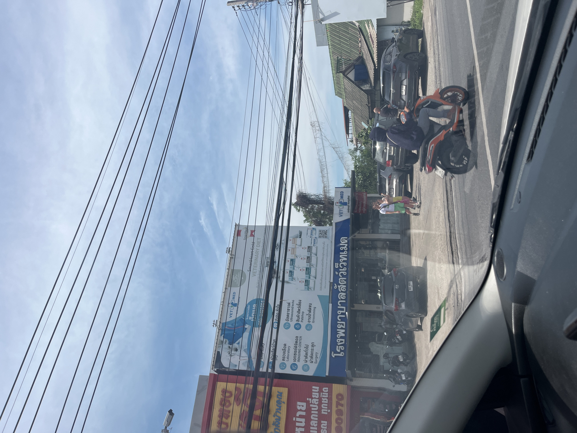 The image depicts a street scene in Rawai, Thailand, on a bright, cloudy day. The foreground shows the interior of a vehicle, with the dashboard and windshield visible. Outside the vehicle, a two-lane road runs horizontally across the frame. On the left side of the road, a veterinary clinic named "VET MED Animal Hospital" is prominently featured, with a large blue and white billboard showcasing various services like laboratory, imaging, and grooming, alongside images of pets and pet food. Below the billboard, the clinic's name is also displayed in Thai script. Adjacent to the clinic is another building with a red facade and Thai text.
Several motorcycles are parked in front of the buildings. A dark grey Mazda hatchback is parked in front of the veterinary clinic. In the middle ground, a person on an orange scooter is riding on the road. Behind the scooter, a dark grey Mazda CX-5 SUV is parked. Further down the street, other vehicles and buildings are visible. Two women and a man are standing on the sidewalk in front of the veterinary clinic. A tangle of electrical wires is strung across the sky overhead. The overall environment appears to be a commercial area in a Thai town.