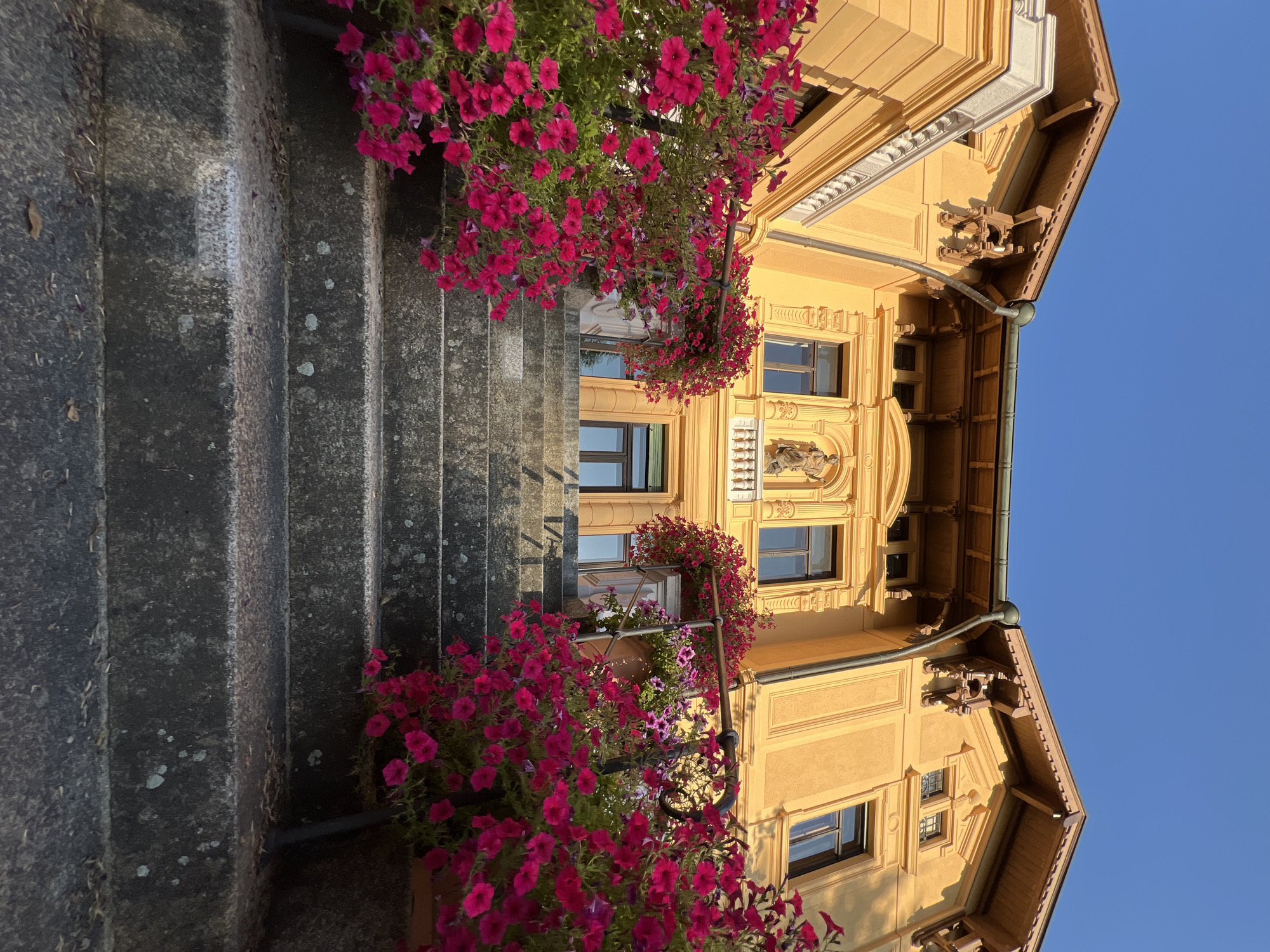 This is a low-angle shot of a yellow building with a detailed facade and a wide stone staircase leading up to it. The staircase is flanked by overflowing planters filled with vibrant pink petunias. The building's architecture features ornate wooden trim under the eaves, decorative sculptures above the windows, and a central balcony with a statue. The sky is a clear, bright blue, suggesting a sunny day. The scene is located in Immenstadt im Allgäu, Germany, specifically at the entrance to the historical hotel "Alte Post."