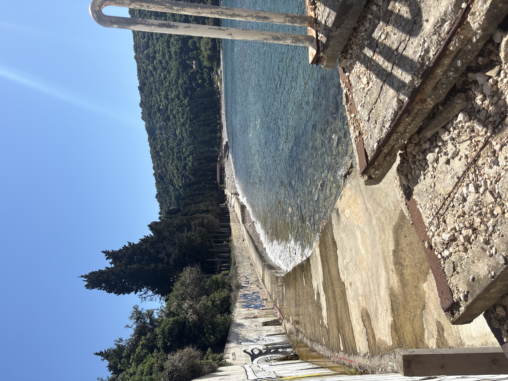The image shows a view of the Adriatic Sea from a concrete pier in Bratica-Braticë, Montenegro. The pier has rusted metal reinforcements and is marked with graffiti. Lush green trees cover the adjacent land. Clear blue sky is visible above. The water is clear and slightly choppy with a small wave breaking on the shore. A rusted metal railing runs along the top edge of the pier.