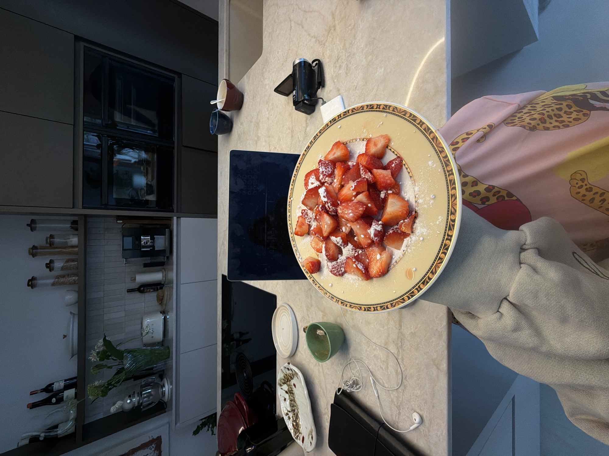 In a modern kitchen, a plate of sliced strawberries dusted with powdered sugar sits on a marble countertop. Next to the plate are white earbuds connected to a black notebook, a small green cup, and an ornate rectangular dish. Further back, a laptop is open, and a small black video camera rests on a white square. Two small bowls, one dark blue and one brown, are positioned on the edge of the counter. In the background, shelves display kitchenware, including bottles of wine, jars, and a coffee machine. The kitchen appears to be located in a home in Ridderkerk, Netherlands.