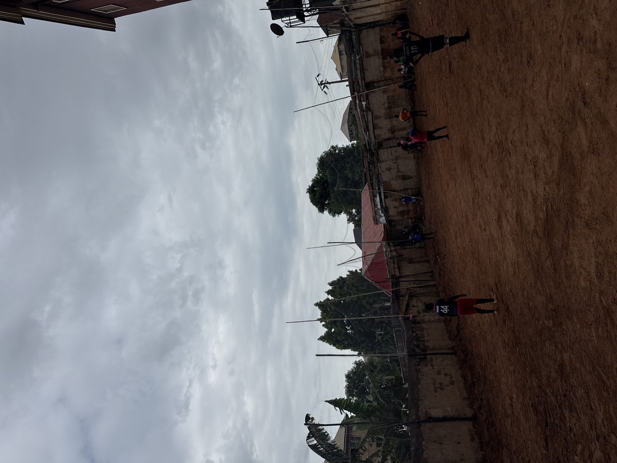 The image depicts an outdoor scene in Nkpor, Nigeria. Several individuals are scattered across a red dirt field. The field is surrounded by a weathered concrete wall topped with a wire mesh fence, with trees and buildings visible behind the fence. Utility poles are also visible. The sky is overcast with grey clouds. Several figures appear to be engaged in activity, though the specifics are unclear. One individual is wearing a dark blue jersey with a red number 44 on the back. The environment is urban and somewhat dilapidated.