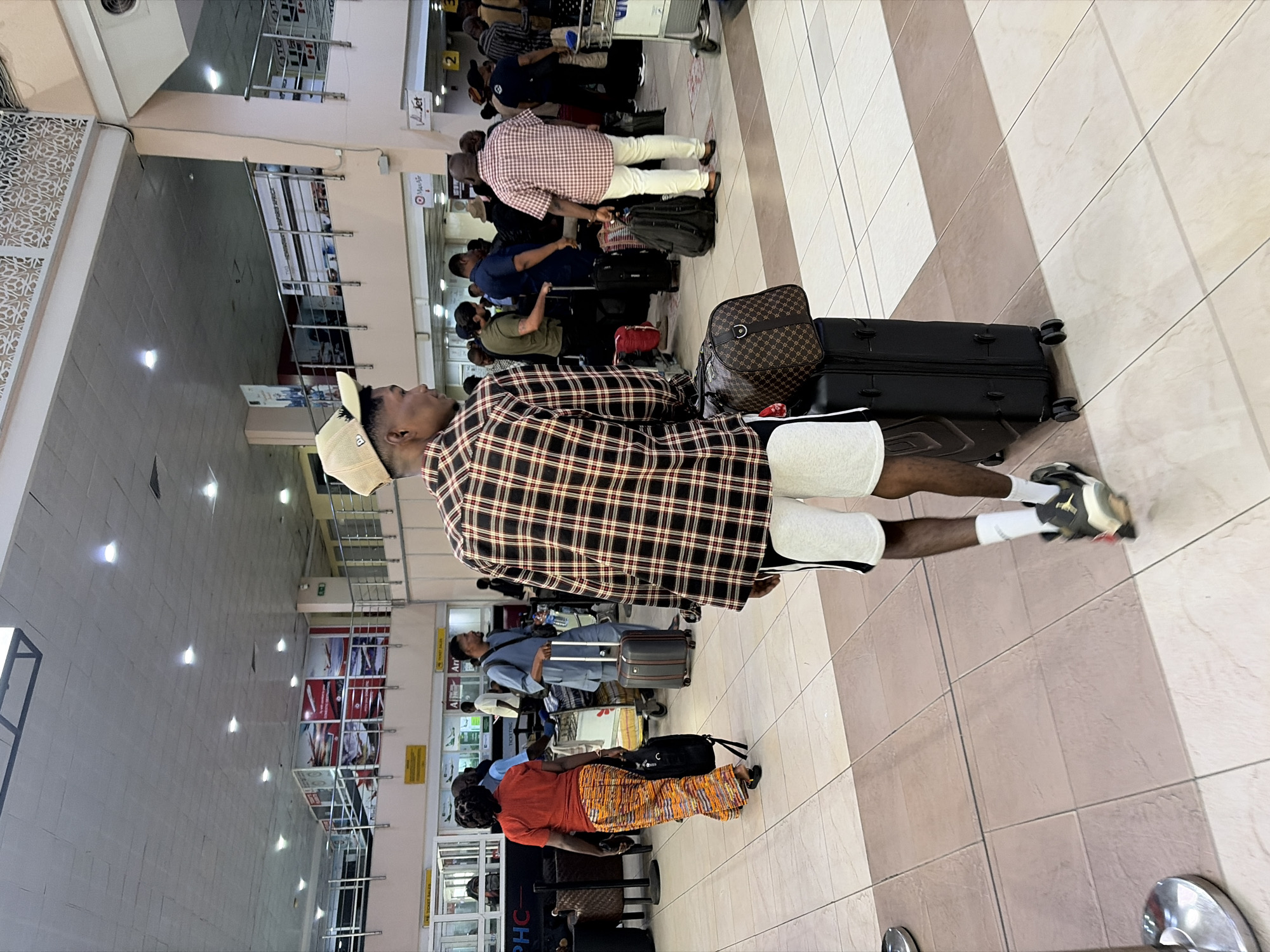 The image depicts a bustling indoor airport terminal environment. The location is identified as being within Ikeja, Nigeria, likely Murtala Muhammed International Airport (MMIA).
In the foreground, a dark-skinned male is viewed from behind, walking right to left, pushing a black, four-wheeled rolling suitcase. A brown, patterned duffel bag with a dark leather trim rests atop the suitcase. He wears a beige baseball cap, a long-sleeved plaid shirt with brown, red, and white checks, light grey shorts with dark side stripes, white socks, and dark sneakers with light soles.
The floor is composed of large rectangular tiles in varying shades of light beige and brown. The ceiling is high, light-colored, and features numerous evenly spaced recessed lights. A mezzanine level with metal railings and advertising banners is visible in the upper portion of the image. One large banner displays images of airplanes, while another, partially obscured, reads "HEARTLAND SAFARIS AFRICA." Decorative white lattice-work is present on an upper wall section.
Throughout the midground and background, numerous individuals, primarily appearing to be of Black ethnicity, are present. Many are standing or walking with luggage, including rolling suitcases, duffel bags, and backpacks. Some individuals appear to be waiting in lines at various counters.
Visible signage includes yellow overhead panels indicating "TICKET SALES" and white signs with "PHC" and "Arr" (likely "Arrivals"). Logos for "ValueJet" and "Max-Air" are also discernable near counters. On the left, a woman wearing an orange top and a multi-patterned skirt walks to the left, carrying a black backpack. To her right, a man in a light blue garment resembling an agbada-style tunic pushes a grey rolling suitcase. A luggage cart is visible on the far right. The overall scene suggests a busy travel hub during daytime hours, with ambient lighting and activity consistent with airport operations.