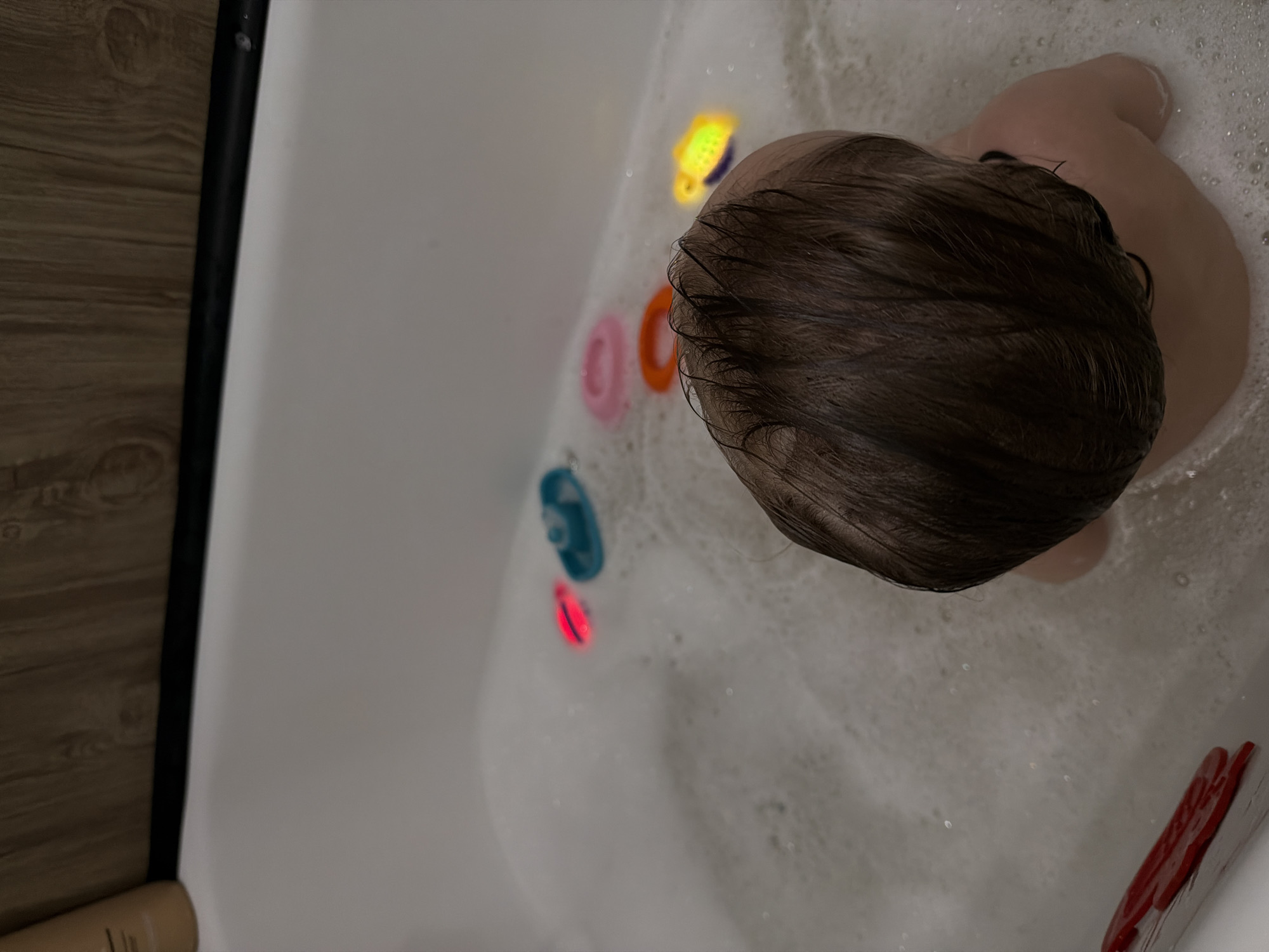 A young child is depicted from an overhead perspective, seated in a white bathtub filled with bubbly water. The child's wet, dark brown hair covers the majority of their visible head, and a portion of their bare back and shoulder are visible, indicating they are immersed in the bath.
Numerous colorful bath toys float around the child. A bright yellow toy, possibly a fish or crab, emits a distinct glow near the child's head. Two plastic rings, one pink and one orange, are also visible. Towards the lower left, a blue toy and a small red glowing object are seen in the water. Another red object, possibly a washcloth or a flat toy, rests at the bottom right edge of the tub.
To the left of the tub, a wooden floor or paneling is visible, separated from the tub by a black vertical rod, possibly part of a shower curtain or frame. A light-colored bottle or container is partially visible on the floor at the bottom left. The glowing toys suggest a dimly lit bathroom, enhancing their luminescence, creating a peaceful and playful atmosphere. No visible text is present. The scene captures a child enjoying a relaxing bath.