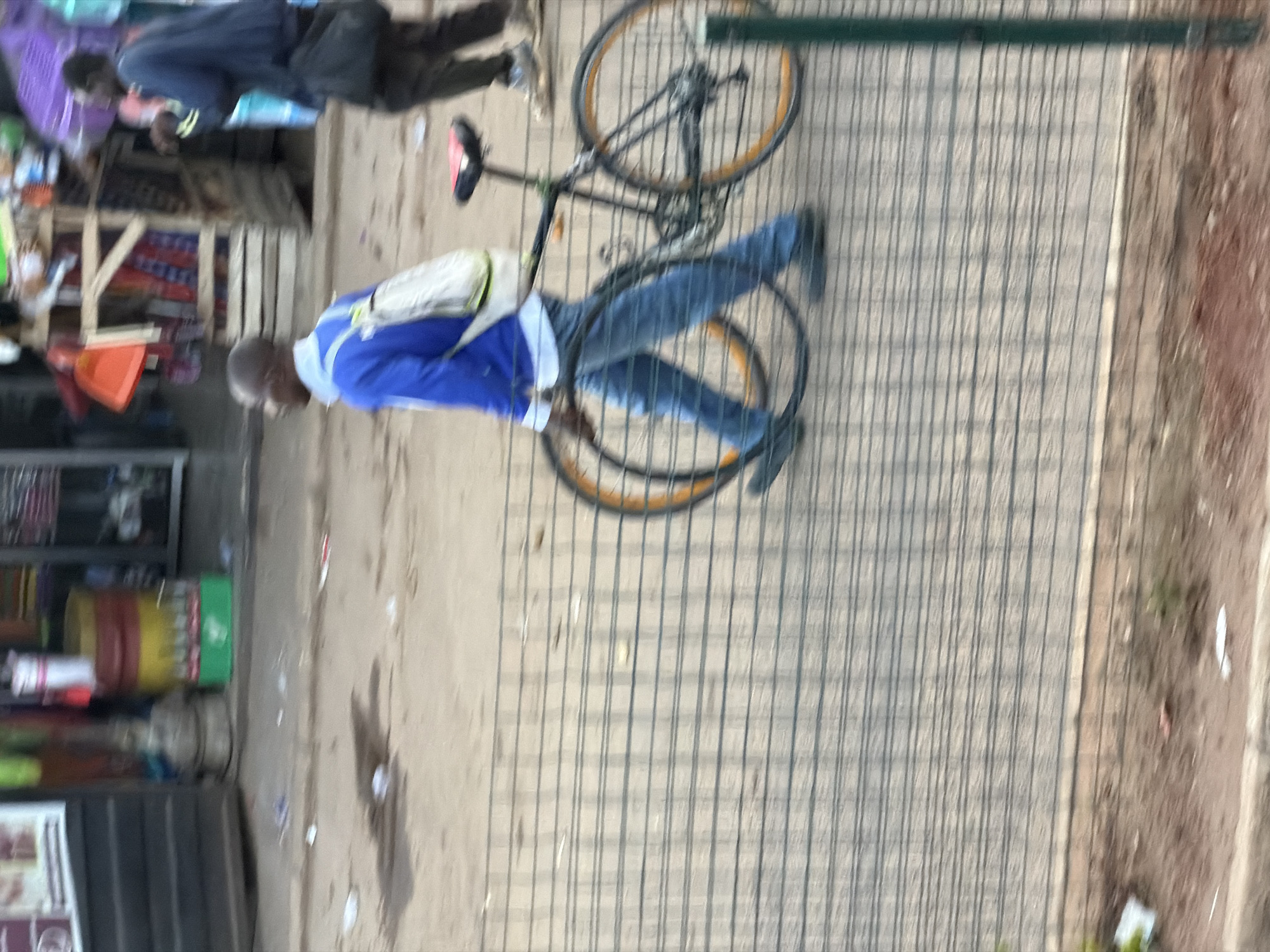 A person wearing a blue jacket and jeans is riding a bicycle on a paved surface. The bicycle has orange rims and a black frame. The person is carrying a white bag on their back. In the background, there are several market stalls with various items for sale, including what appear to be food and drinks. The scene suggests a busy marketplace in Bukuru, Nigeria. The lighting indicates it is daytime, but the weather is not discernible. There is no visible text in the image.