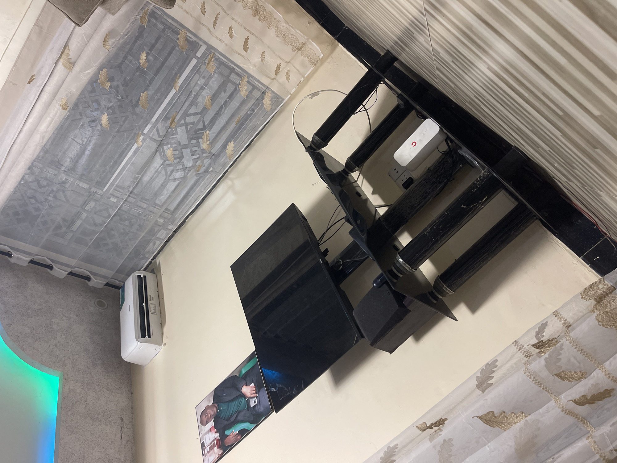 A residential room in Dar es-Salaam, Tanzania, exhibits a TV mounted on a wall-mounted black metal rack. Below the TV, a framed picture of a man in a green shirt is displayed. An air conditioning unit is affixed to the wall. Curtains with gold leaf patterns are hung in front of the windows. A white router is positioned on the top shelf of the metal rack. The walls are painted light beige. A section of a wall with green and blue LED lighting is visible on the left.