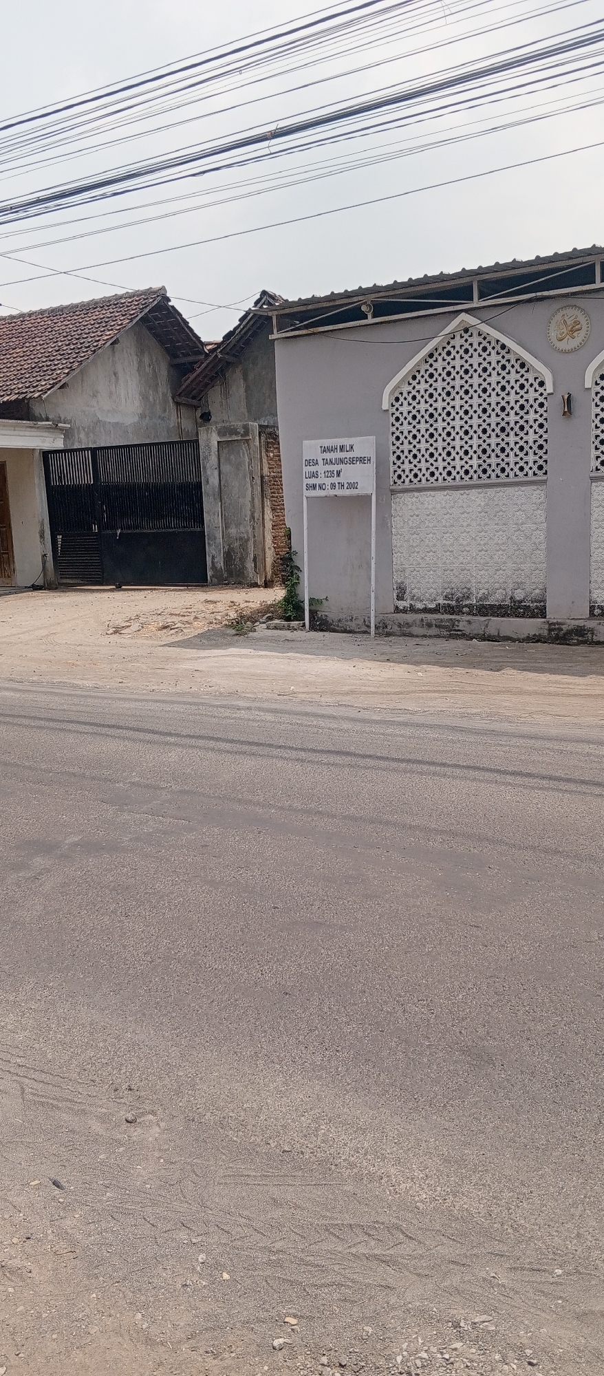 The image depicts a street scene in Gulun, Indonesia, with a focus on a property boundary sign. In the foreground, a paved road occupies the lower half of the frame, showing tire tracks and scattered gravel. The middle ground reveals a grey building with decorative white latticework and a crescent moon emblem with Arabic script. Adjacent to this building is a white signpost. The sign itself is rectangular and white, with black text. The text on the sign reads: "TANAH MILIK DESA TANJUNGSEPREH LUAS : 1235 M² SHM NO : 09 TH 2002". This translates to "PROPERTY OF THE VILLAGE OF TANJUNGSEPREH AREA: 1235 M² Land Title Certificate NO: 09 YEAR 2002". To the left of the sign and the grey building, a darker, older building with a tiled roof is partially visible, featuring a large black metal gate. The background is dominated by a hazy sky and a network of overhead power lines. The overall scene suggests an area with both residential and possibly communal or religious structures. There are no people visible in the image.