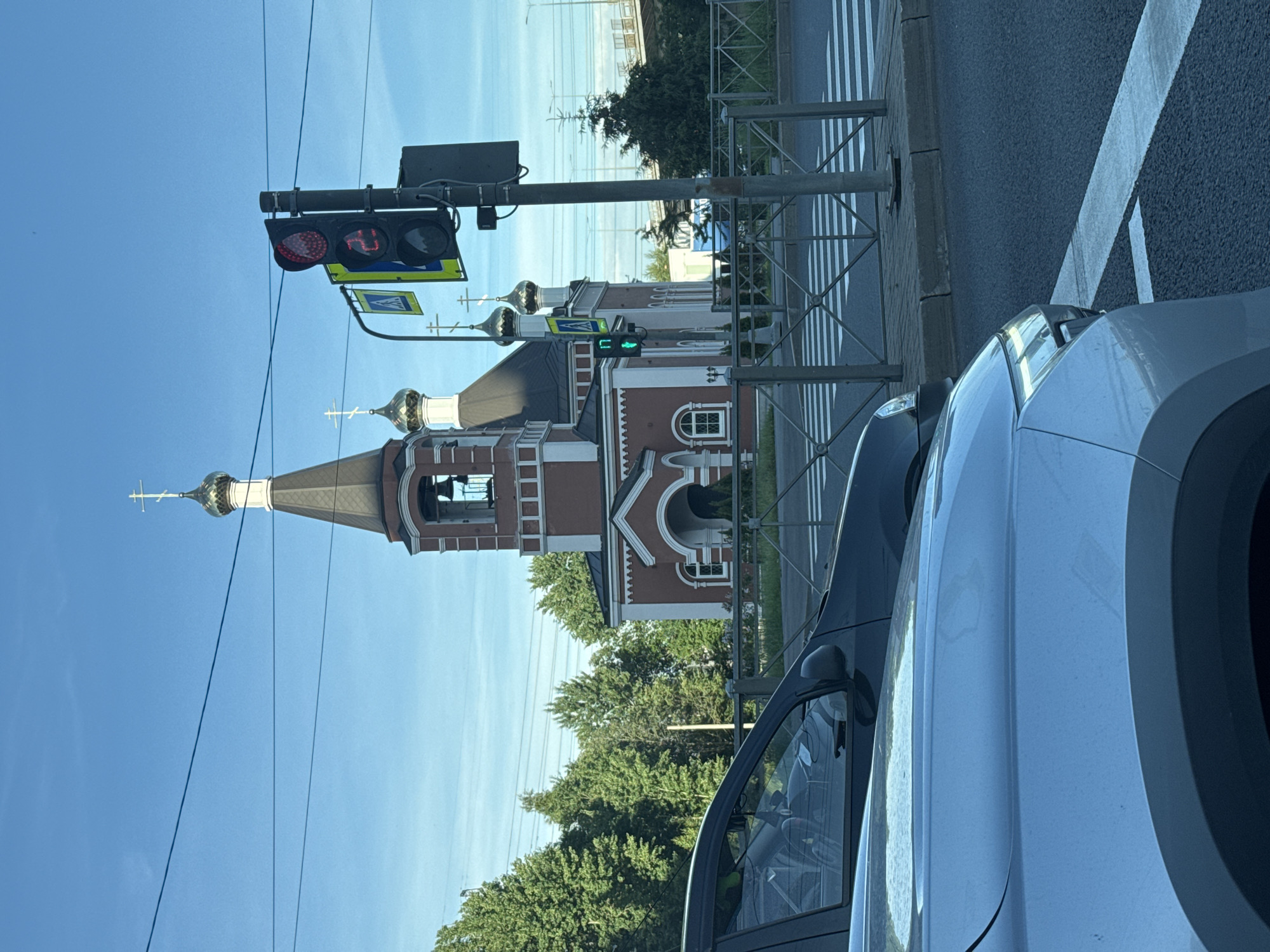 The image shows an Orthodox church with a red brick facade and gold domes against a clear blue sky. In the foreground, two cars are partially visible, one silver and one dark gray. A traffic light is also visible, showing a red light for vehicles and a green walking man symbol for pedestrians. A crosswalk is marked on the road. Trees line the background, with power lines overhead. The scene appears to be an intersection in a city, though the specific location is unknown.