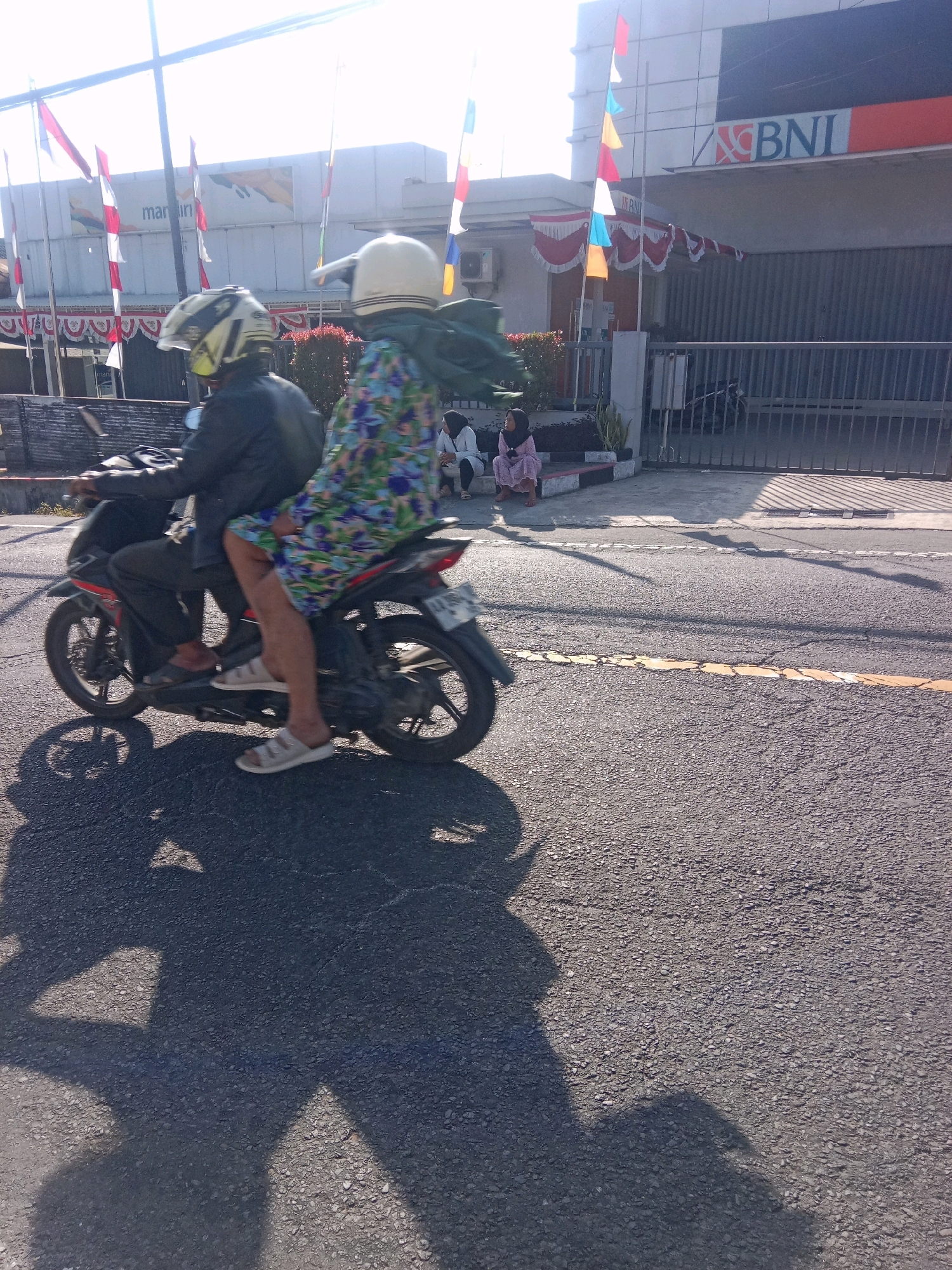 Two individuals are riding a black motorcycle on an asphalt road in front of a building. The person in the front is wearing a black jacket and a white helmet, while the person in the back is wearing a colorful patterned dress and a white helmet. The building behind them appears to be a bank, with a BNI logo visible. There are flags displayed outside the building, and a fence in front of it. The road has yellow lane markings. The scene is illuminated by sunlight, casting shadows of the motorcycle and riders onto the road.