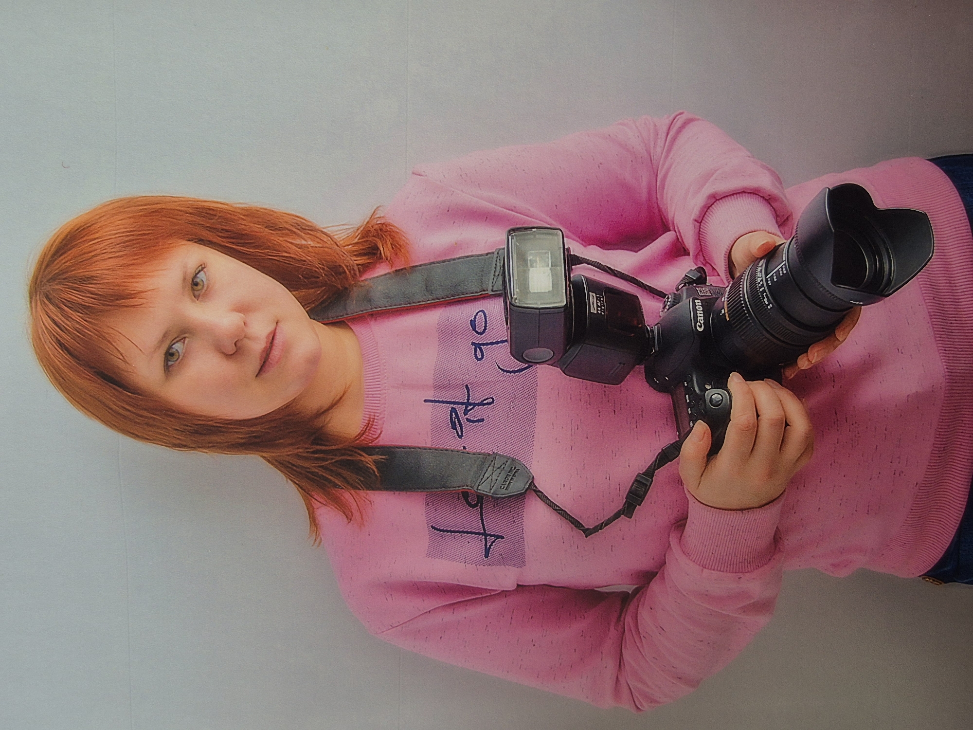 A young woman with reddish-orange hair and bangs stands against a light gray wall, holding a professional camera with a flash. She is wearing a pink sweatshirt with a graphic design across the chest and a dark gray camera strap around her neck. The woman is looking directly at the camera with a neutral expression. Her hands are positioned on the camera and lens, suggesting she is either about to take a photo or has just finished. The lighting is soft, creating a slight shadow on the wall behind her. There are no other people or objects visible in the frame. The image is a medium shot, focusing on the woman and her camera.