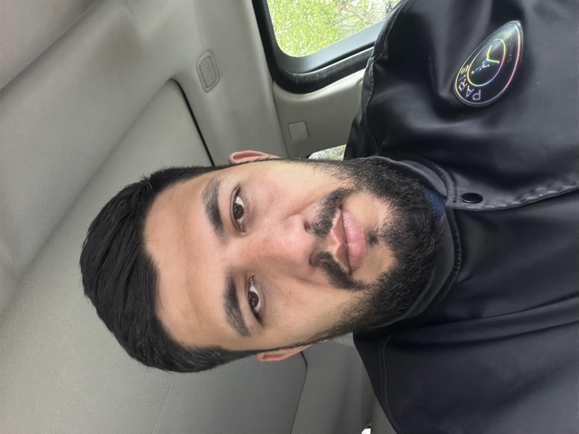A man with dark hair and a beard is sitting in a car. He is wearing a black jacket with a "Paris Saint Germain" logo on the right side. The interior of the car is visible, with a light grey ceiling and part of a window.
It is not possible to determine the location as Krasnoyarsk, Russia from the provided image.