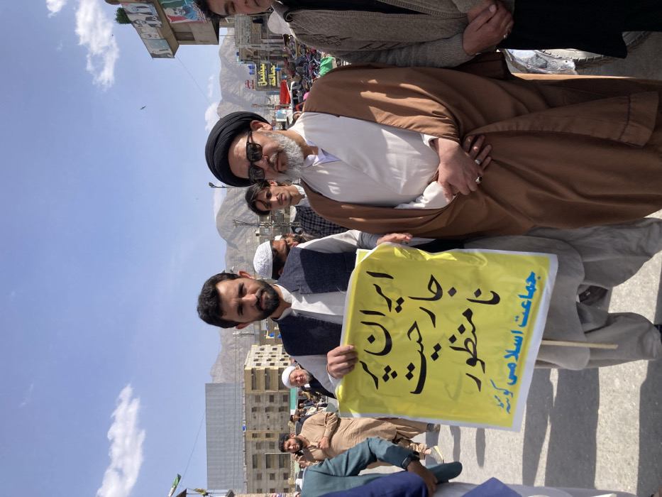 The image depicts an outdoor daytime scene in Quetta City Tehsil, Pakistan, showing a gathering of men. In the foreground, two men are prominent. The man on the left, with a dark beard and wearing a white shalwar kameez with a dark waistcoat, holds a yellow placard. The placard features black Urdu script reading, "ایران پر جارحیت نامنظور" (Aggression against Iran is unacceptable) and "جماعت اسلامی کوئٹہ" (Jamaat-e-Islami Quetta). He looks directly at the camera. To his right, another man, wearing sunglasses, a black turban, and a brown cloak over a white tunic, stands with his hands clasped in front of him, looking slightly to his right.
Behind these two individuals, numerous other men are visible, dressed in various forms of traditional attire, including shalwar kameez and prayer caps. The background features multi-story buildings lining a street or public area. Some buildings display commercial signage, including one with "سپیشل" (Special) in Urdu and another with political banners. Distant mountains are visible under a light blue sky with scattered white clouds. The ground appears paved.