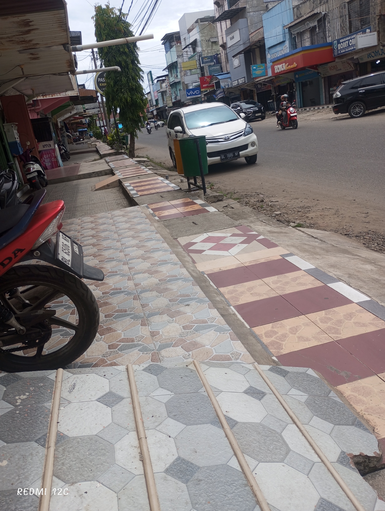 This image shows a street scene in Lhokseumawe, Indonesia. The foreground features a tiled sidewalk with three metal bars embedded in it. A motorcycle is parked on the sidewalk. In the street, a white minivan with the license plate BL 1686 NF is driving. A red scooter is also traveling in the same direction as the van. Buildings line the street, with signage visible for businesses like Kodak, Dulux, Roma, Vivo, Money Changer, Quick Laundry, and Pas Express. The overall scene appears to be a busy commercial area in Lhokseumawe. The weather appears to be overcast.