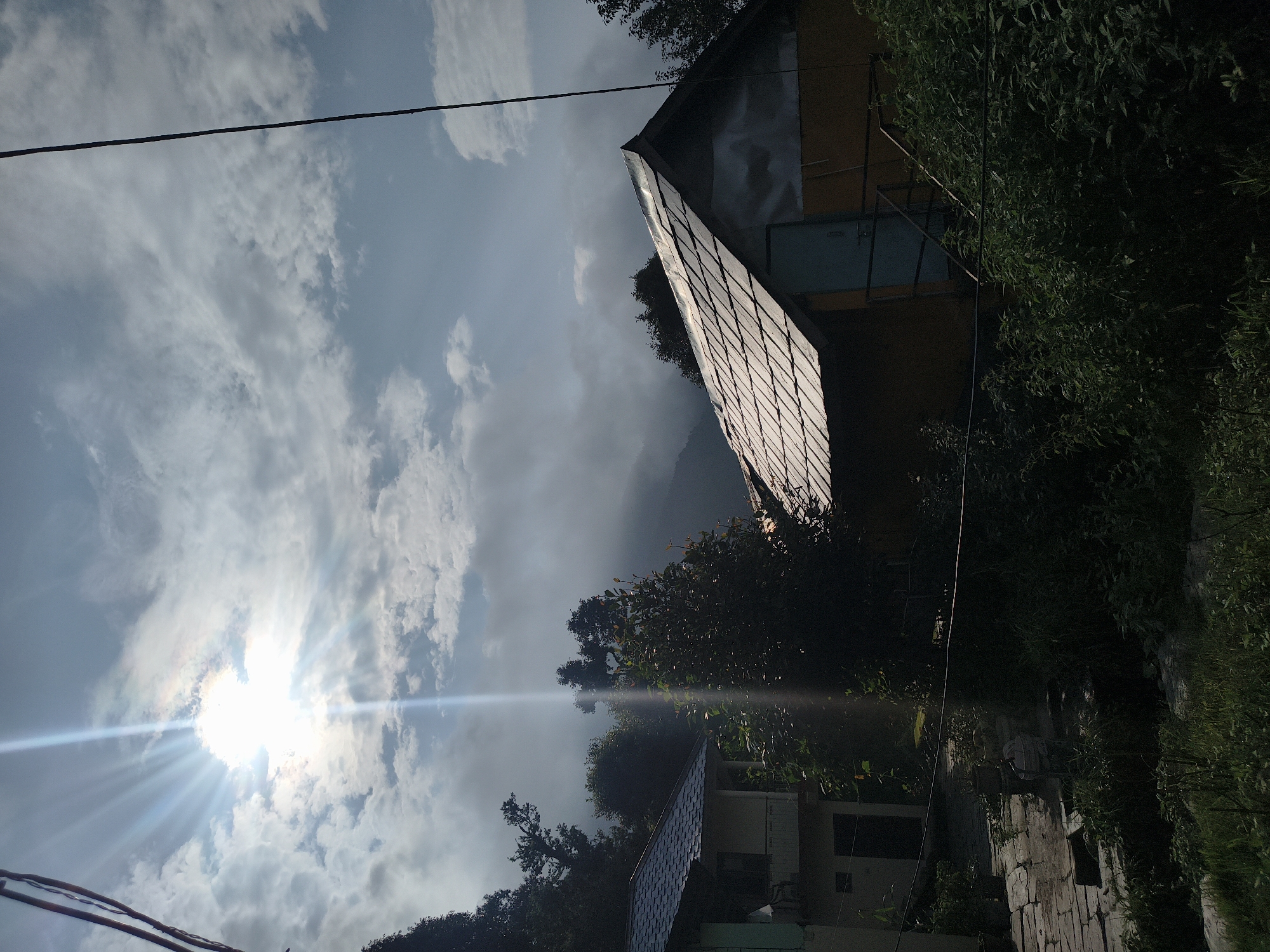 The photograph depicts a rural scene in Haini, India, dominated by a dramatic sky and a rustic dwelling. The sun, a brilliant white orb, shines through a cloudy sky, casting strong rays that create a lens flare effect across the upper half of the image. Wispy clouds drift across the blue sky, some appearing as dark silhouettes against the brighter sky. In the mid-ground, a traditional-style house with a gray, tiled roof is visible. The house appears to be made of a yellow material and has a light blue door. A metal staircase leads to this door. To the left of this house, another building with a light-colored facade and a blue tiled roof is partially visible. Lush green foliage, including trees and bushes, fills the foreground and surrounds the structures, suggesting a natural, overgrown environment. The overall impression is one of a tranquil, perhaps remote, village setting under a dynamic sky.
