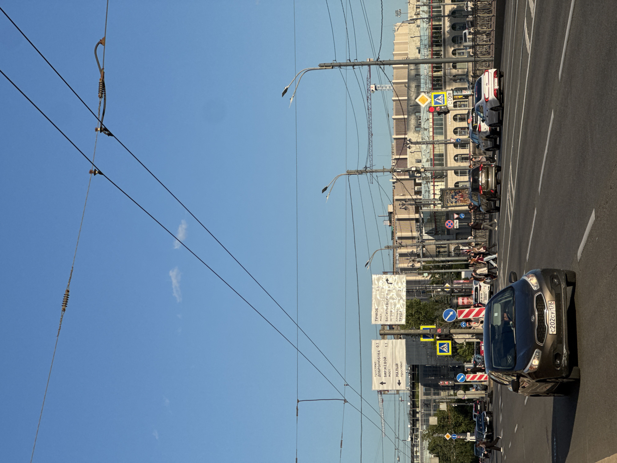 The image depicts a street scene in Saint Petersburg, Russia, likely a bridge, with a clear blue sky and overhead wires. In the foreground, a dark-colored Kia car drives on the right side of the road, displaying a Russian license plate: "C202CY 178". Several other vehicles are visible further down the road, including an SUV and a white sedan. Pedestrians are crossing the street at a crosswalk, with traffic lights visible. Directional signs are mounted on poles, indicating distances to locations such as "prospekt Dobrolyubova" (Dobrolyubova Avenue), "Birzhevoy most" (Birzhevoy Bridge), and "Malyl prospekt" (Small Avenue). In the background, a large, modern building with multiple windows and arched entrances dominates the skyline, with a crane visible to its left. The overall scene suggests an active urban environment.