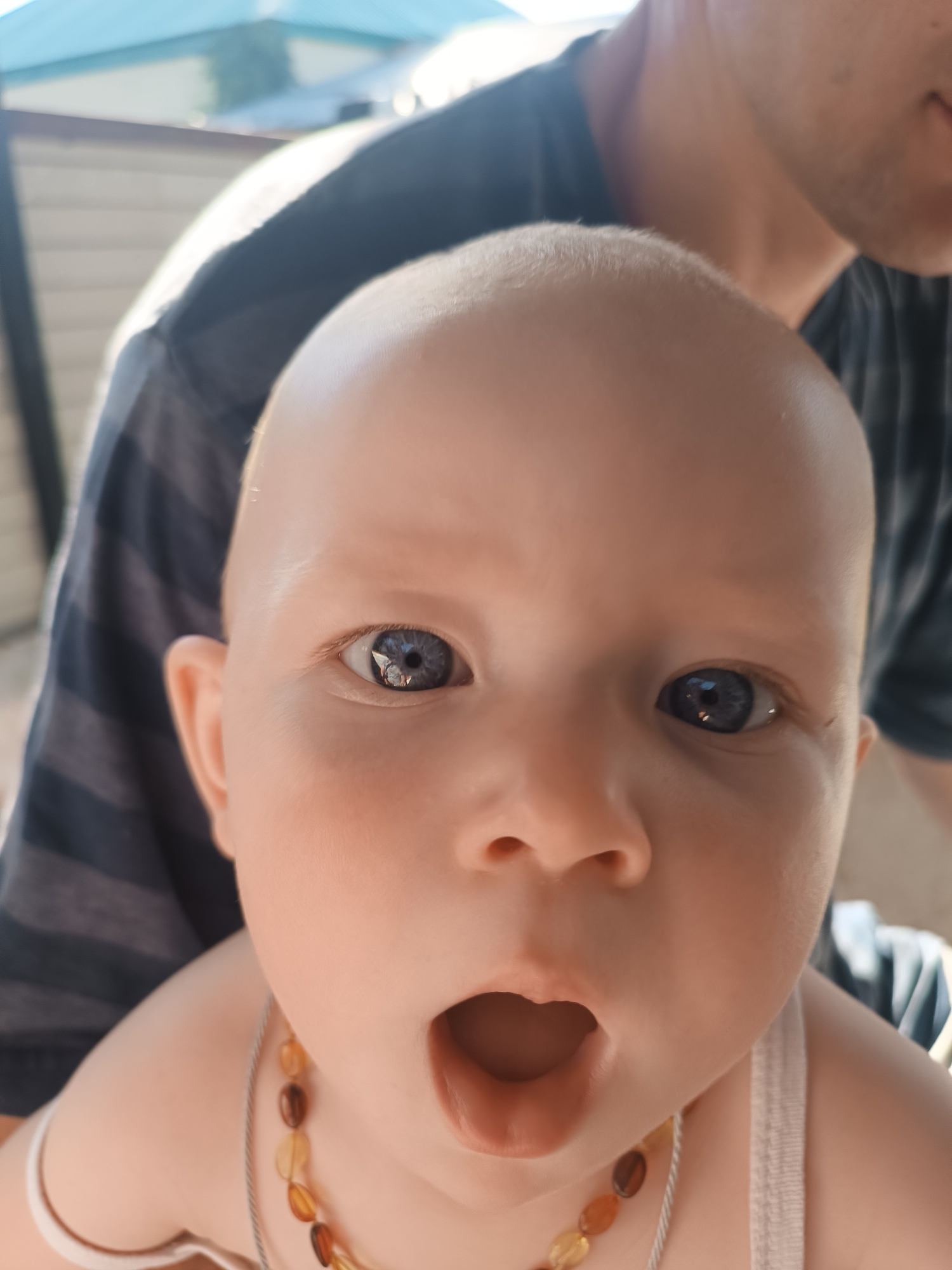 A close-up, high-angle outdoor shot features a baby with light skin and blue eyes, and is being held by an adult whose torso is visible. The baby is looking directly at the camera with a surprised expression, mouth agape. The baby is wearing a necklace of amber beads and a white tank top. The adult is wearing a dark t-shirt with horizontal stripes. The background shows a blurred view of outdoor structures and foliage, suggesting a residential area. The scene is lit by natural sunlight, casting soft shadows and highlights.