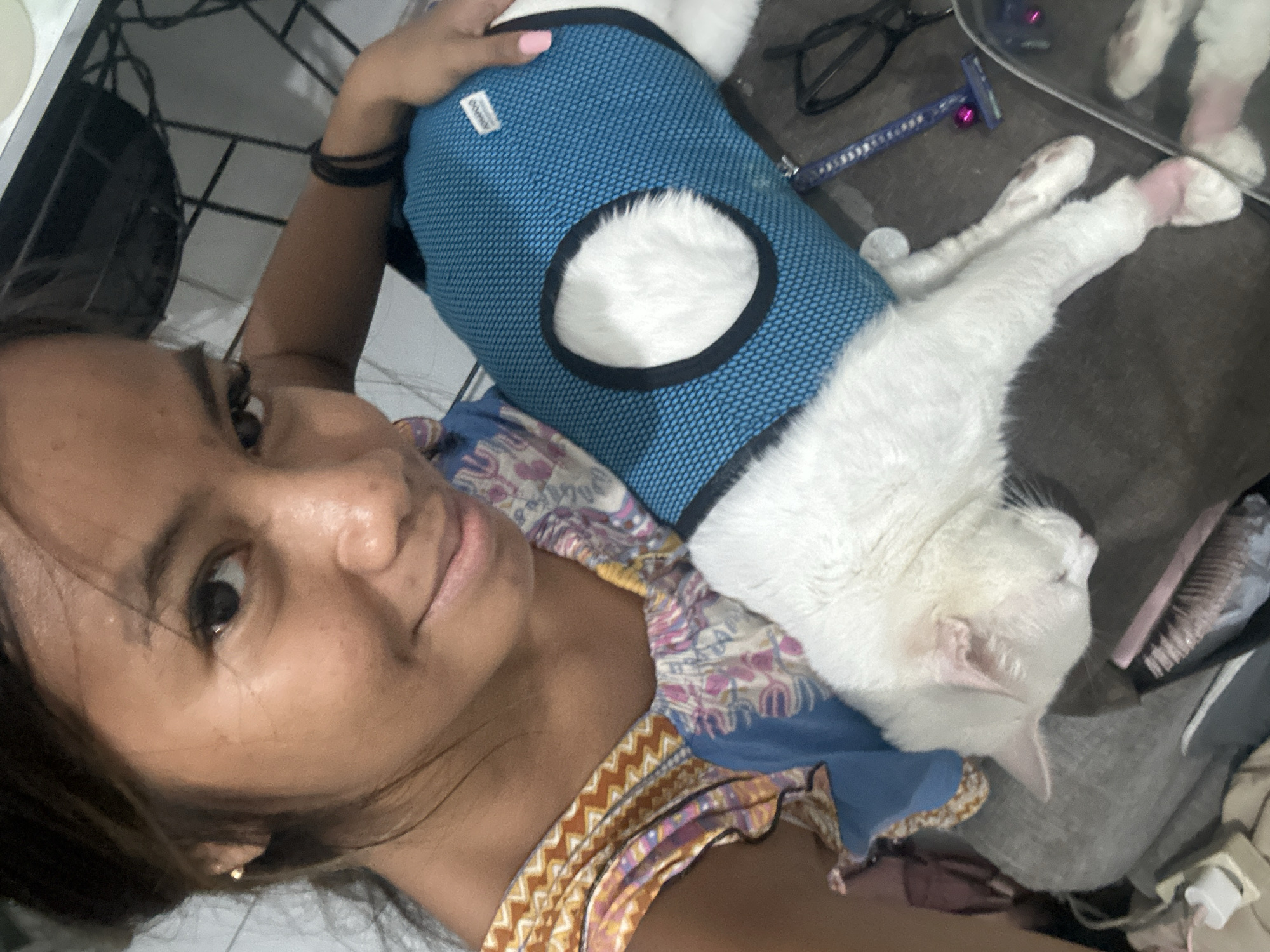 A woman poses for a selfie, cradling a white cat wearing a blue vest. The cat is relaxed and resting on the woman. The background includes a pair of glasses, a disposable razor, and miscellaneous items on a surface that appears to be a cluttered floor or bed. No specific location information in North Jakarta, Indonesia, can be determined from the image alone.