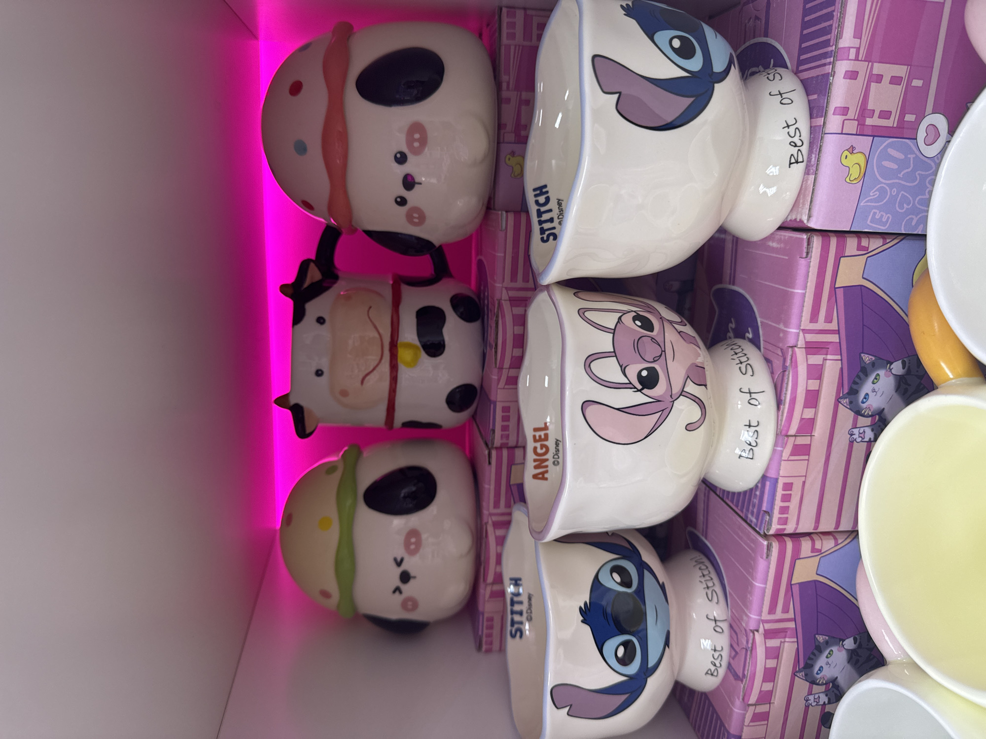 The image shows an assortment of novelty mugs and their packaging, displayed under a pink light source. On the left, there are three stacked character mugs. The top mug features a white character face with pink accents, the middle one resembles a cow, and the bottom one is another white character face with green accents.
To the right of the mugs are a set of character-themed bowls, each in its respective packaging. The designs include "Stitch" and "Angel" from Disney. The packaging has a lilac-themed design, featuring cartoon drawings. The "Stitch" bowls are marked with "Best of Stitch!".
This merchandise is likely being sold or displayed at a retail location. There is no concrete evidence that the location is Chaikovsky, Russia.