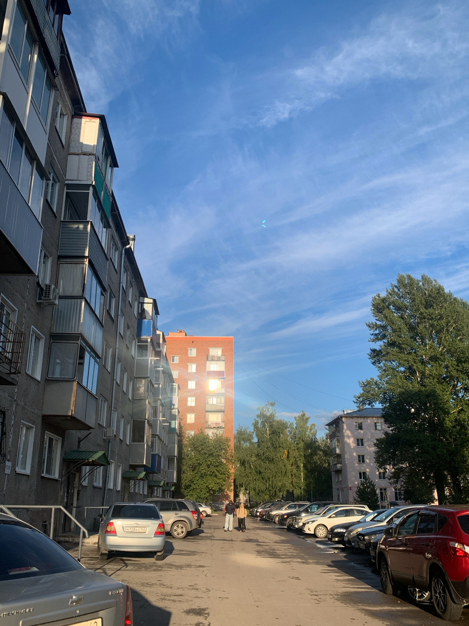 This is an eye-level, outdoor shot of a residential street in Prokopyevsk, Russia, during the daytime. The scene is dominated by several apartment buildings, a parking lot filled with cars, and a clear blue sky with wispy clouds.
On the left side of the image, a tall, grey apartment building with numerous windows and balconies stretches along the street. Some balconies are enclosed, while others are open, with some featuring green awnings. An air conditioning unit is visible on the building's facade.
Further down the street, a mid-rise, orange brick apartment building stands out, with the sun reflecting brightly off its windows. Tall, green trees are interspersed between the buildings.
A long row of parked cars fills the asphalt lot. Visible vehicles include a silver sedan with a license plate beginning with "M728AO 142", a silver SUV, a white hatchback, and a red SUV at the front right.
Two individuals are walking away from the camera on the sidewalk in the middle ground. One is wearing a light-colored jacket and dark pants, and the other is in a dark jacket and pants.
In the background, a lighter pink apartment building can be seen, partially obscured by trees. Power lines are also visible. The overall environment suggests a typical urban residential area in Prokopyevsk.