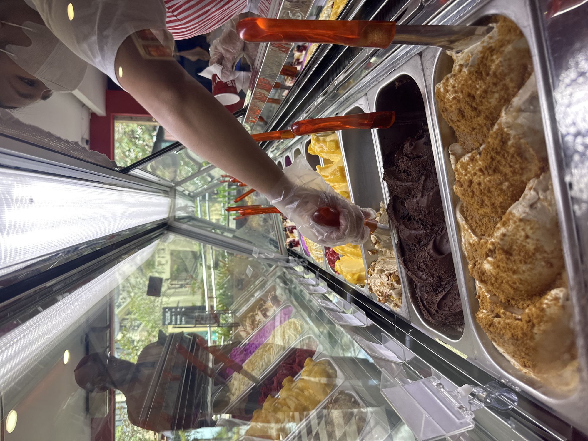 In Kuta Selatan, Indonesia, at a gelato shop, a person wearing a white shirt and red and white striped apron, as well as a face mask, is serving gelato. They are wearing plastic gloves and are using a gelato scoop to serve a customer from a display case filled with various flavors of gelato. The display case is illuminated by overhead lights, reflecting the surrounding environment which includes trees and buildings visible through the glass. In the foreground, several tubs of gelato are visible, including a dark chocolate flavor, and a light brown flavor topped with crumbled cookies. The person is in the process of scooping gelato from a tub of bright yellow gelato.