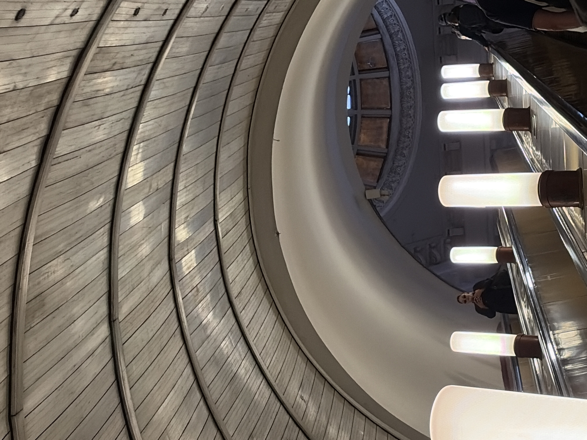 This is a medium shot, eye-level photograph taken inside a subway station. The focus is on the interior architecture and the escalator. The ceiling is a large, curved structure made of wooden planks, forming concentric arches. The walls of the station are white, curving upwards to meet the ceiling. In the center of the image, a large, arched opening reveals a circular dome-like structure with a grid of windows.
On the right side of the image, an escalator is visible, ascending. Several people are on the escalator, dressed in casual clothing. The foremost person on the right is wearing a dark jacket and a baseball cap. Further up the escalator, a person in a black jacket and sunglasses is holding a phone. There are several cylindrical light fixtures along the side of the escalator, emitting a bright, white light. The escalator handrail and steps appear to be made of polished metal. The overall impression is one of a grand, somewhat retro, subway station.